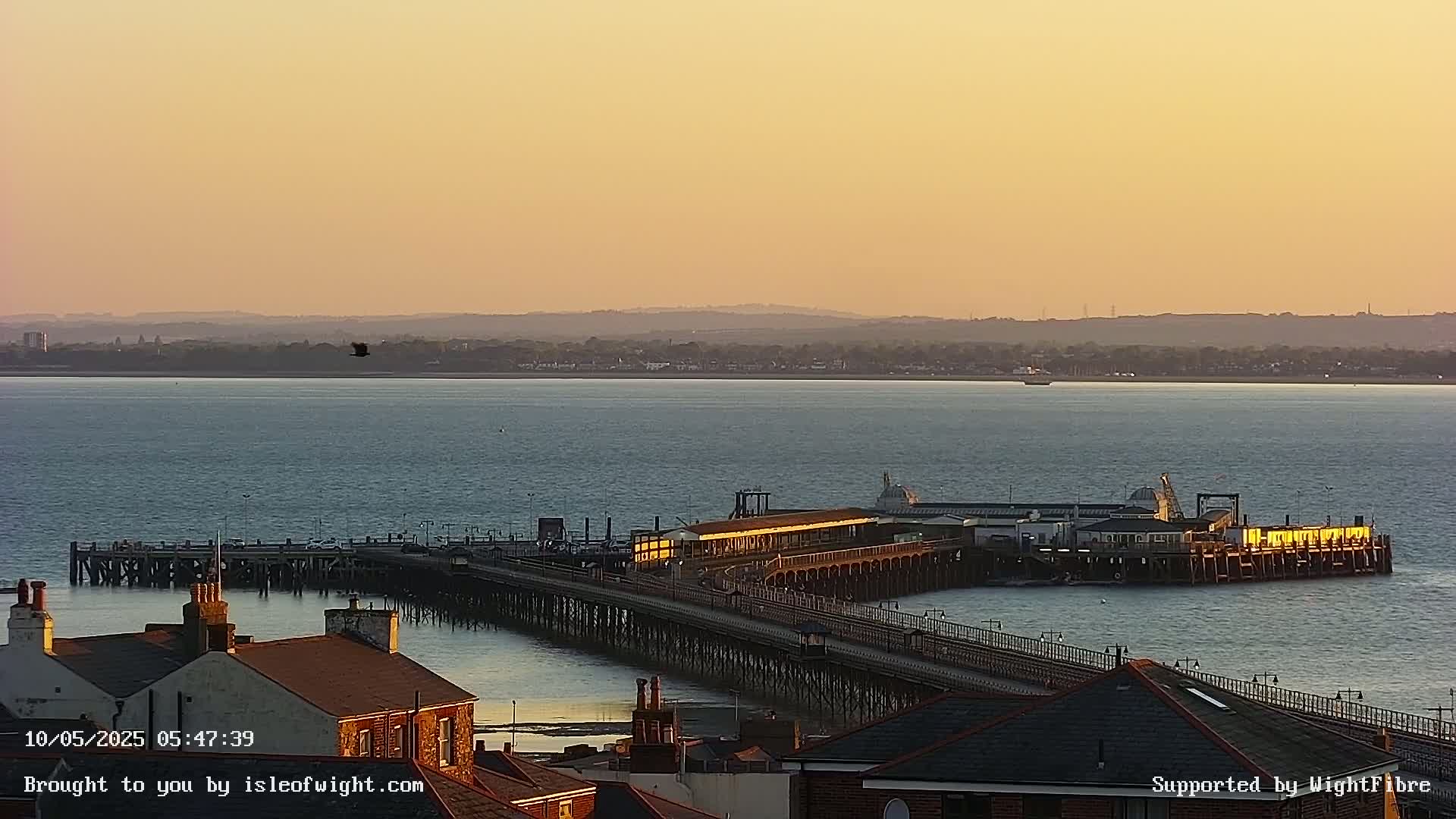 A long pier extends into a calm body of water under a pale orange sunset, with rooftops of buildings visible in the foreground.