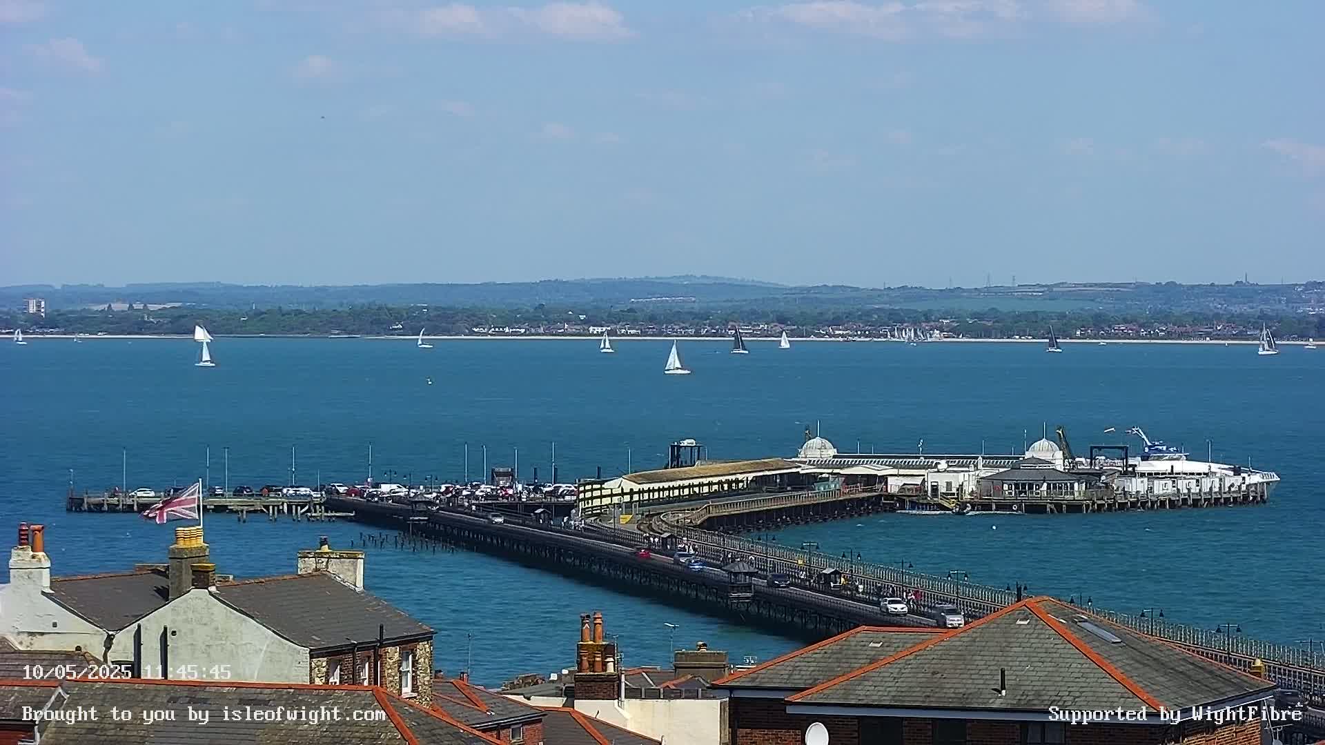 A long pier extends from a town into a calm, blue body of water dotted with sailboats under a clear sky.