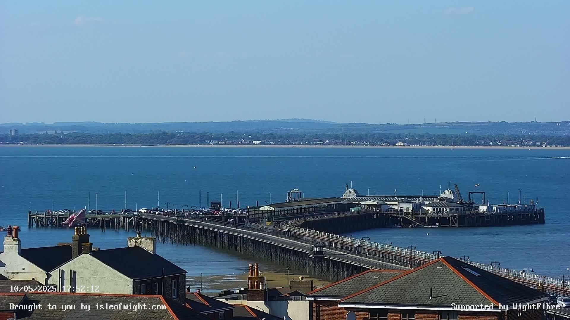 A long pier extends into a calm, blue body of water on a sunny day, with a distant shoreline visible in the background.