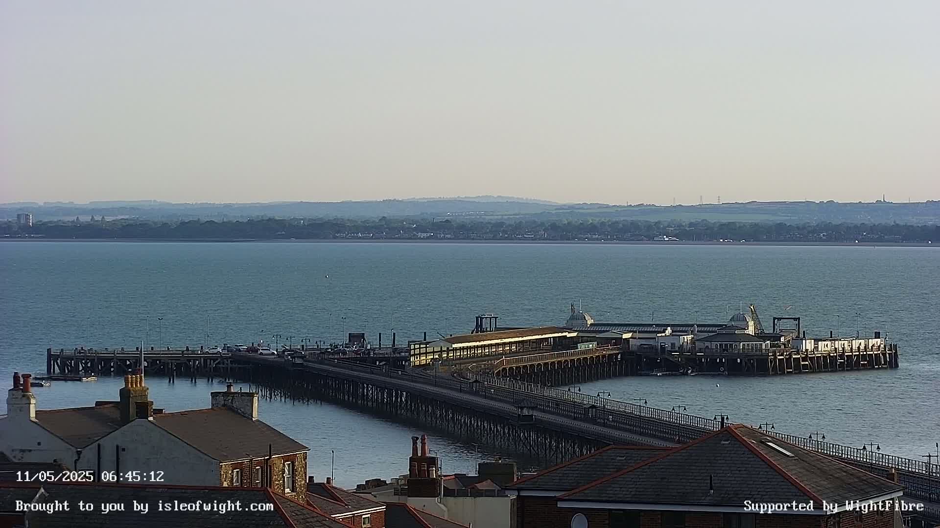 A long pier extends into a calm body of water, with buildings and rooftops visible in the foreground under a hazy sky.