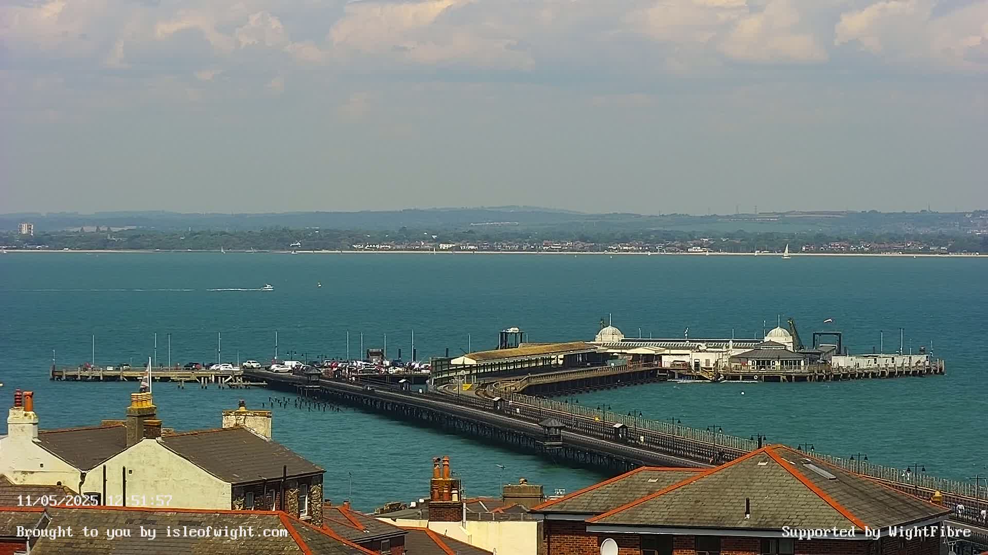 A long pier extends into a calm, teal sea under a partly cloudy sky, with rooftops of buildings visible in the foreground.