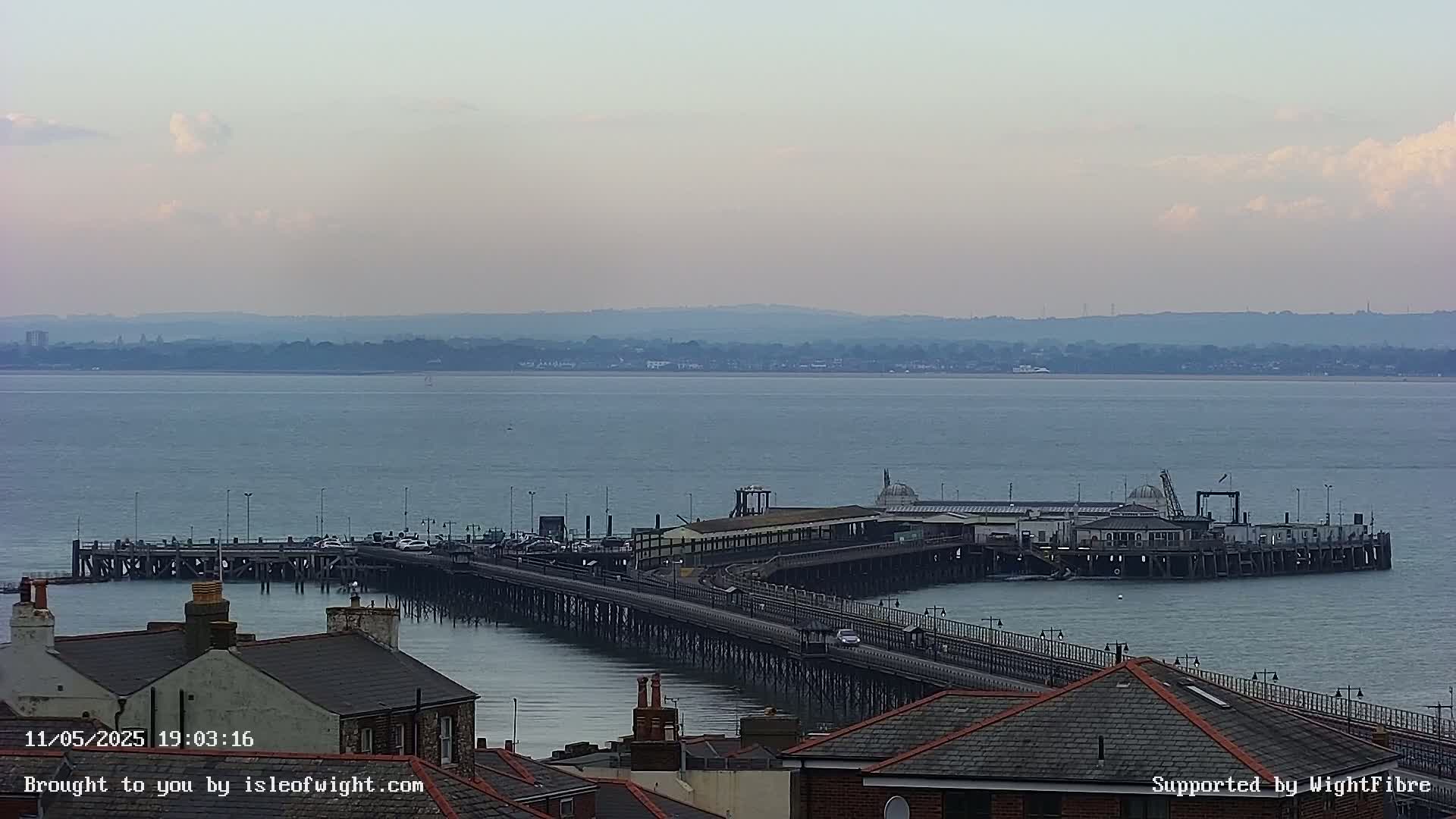 A long pier extends into a calm body of water under a partly cloudy sky, with distant land visible on the horizon.