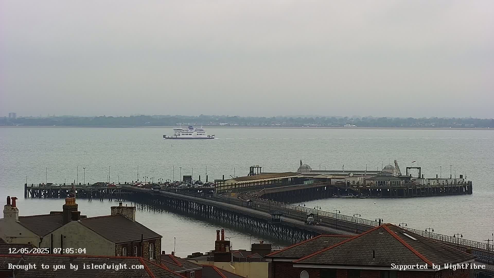 A ferry is seen in the distance on a calm, overcast day, approaching a long pier with buildings and vehicles on it, viewed from a high vantage point overlooking rooftops.
