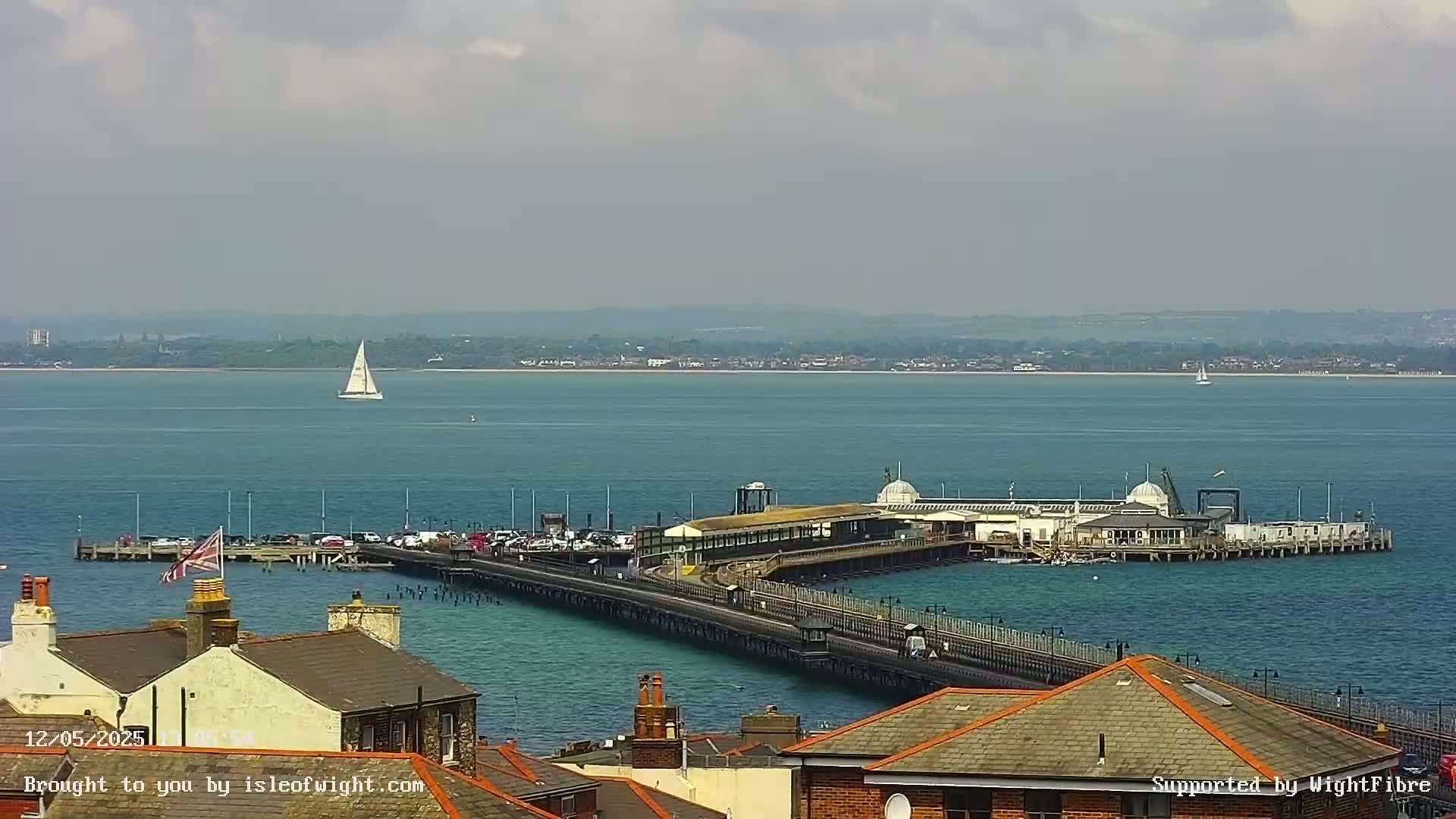 A long pier extends into a calm, blue body of water under a partly cloudy sky, with a sailboat visible in the distance and rooftops in the foreground.
