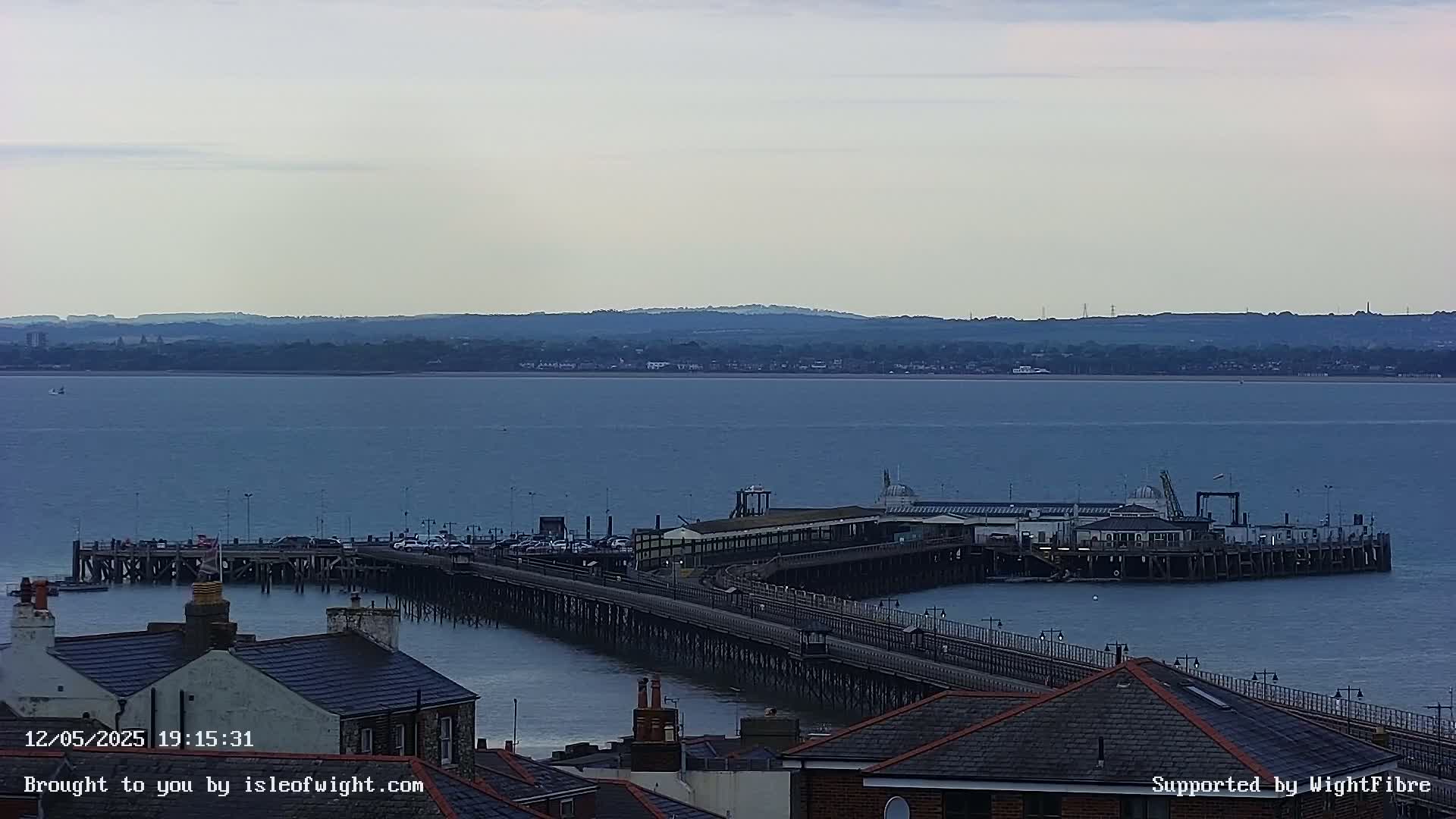 A long pier extends from a shoreline into a calm, blue body of water, with a distant landmass visible under a cloudy sky.