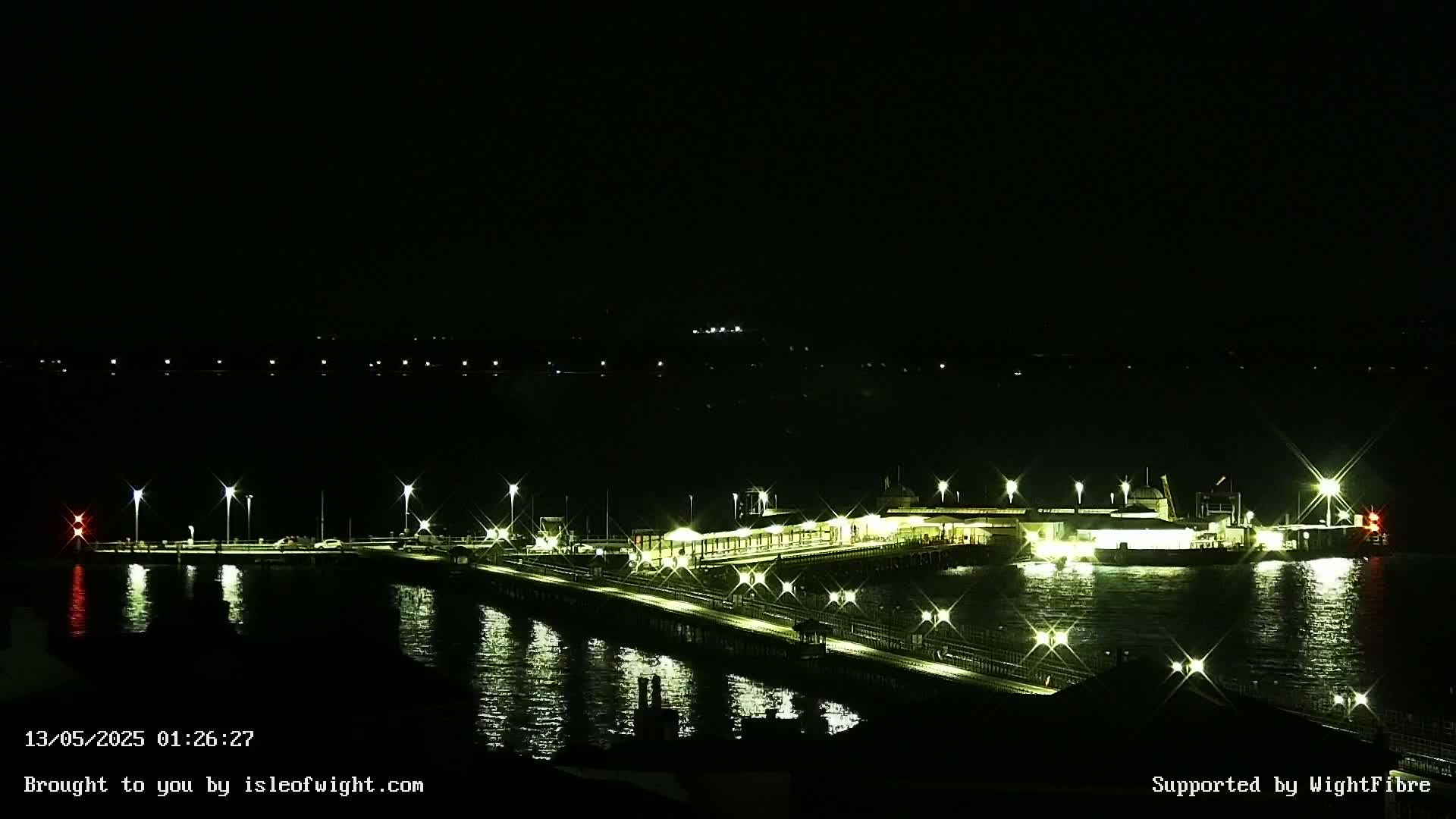 A brightly lit ferry terminal and pier are reflected in dark, calm water under a nighttime sky.