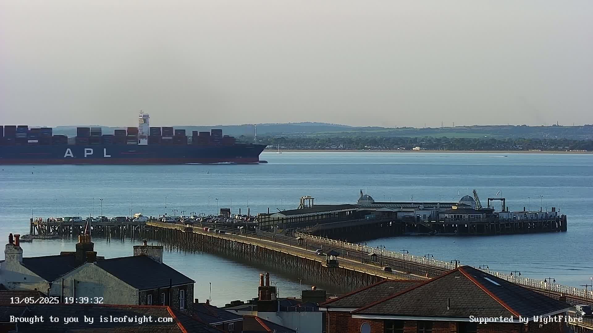 A large container ship is visible in the distance across calm water from a long pier with buildings and vehicles on it.