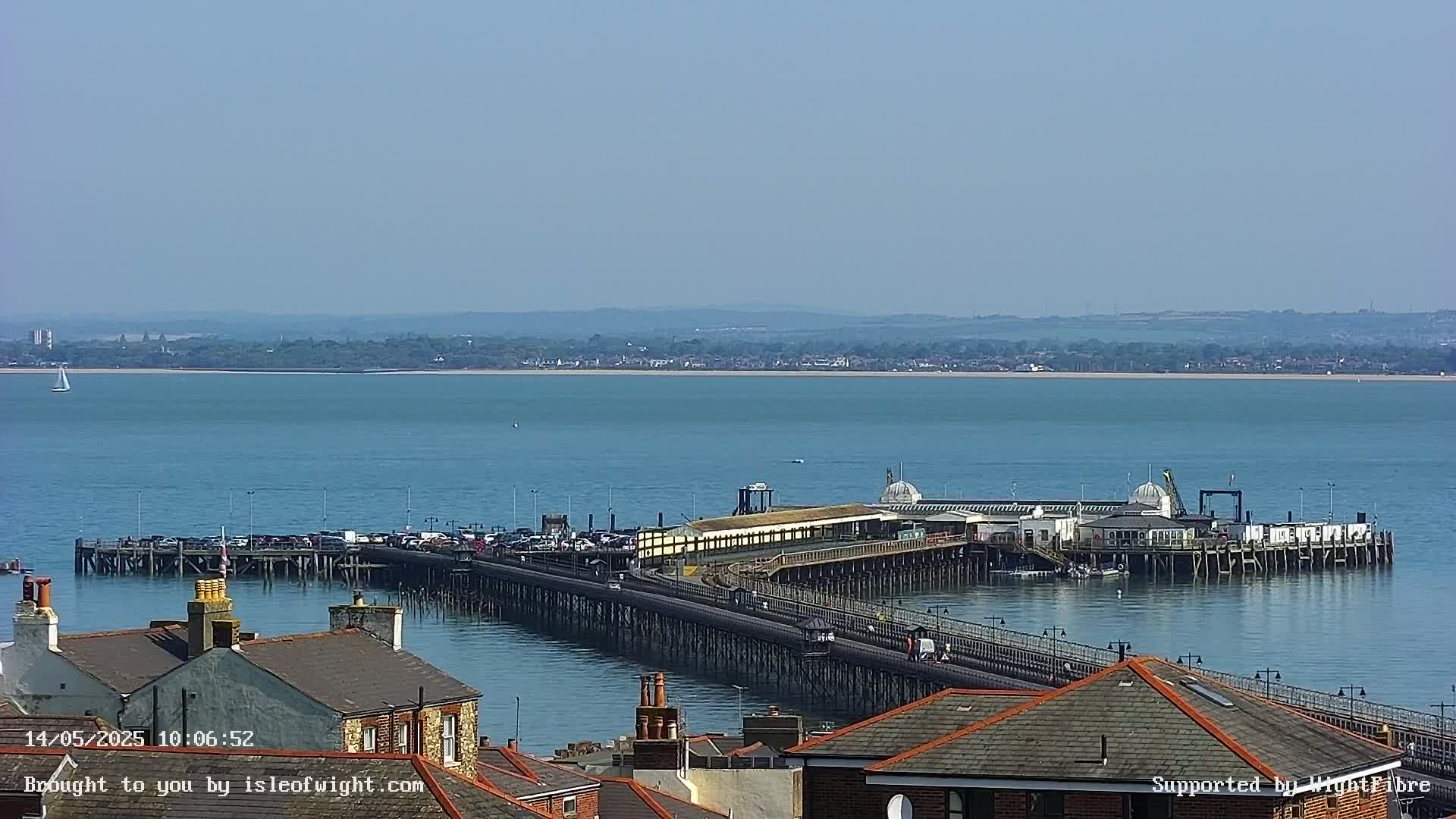 A long pier extends into a calm, blue body of water on a sunny day, with rooftops and a distant shoreline visible.