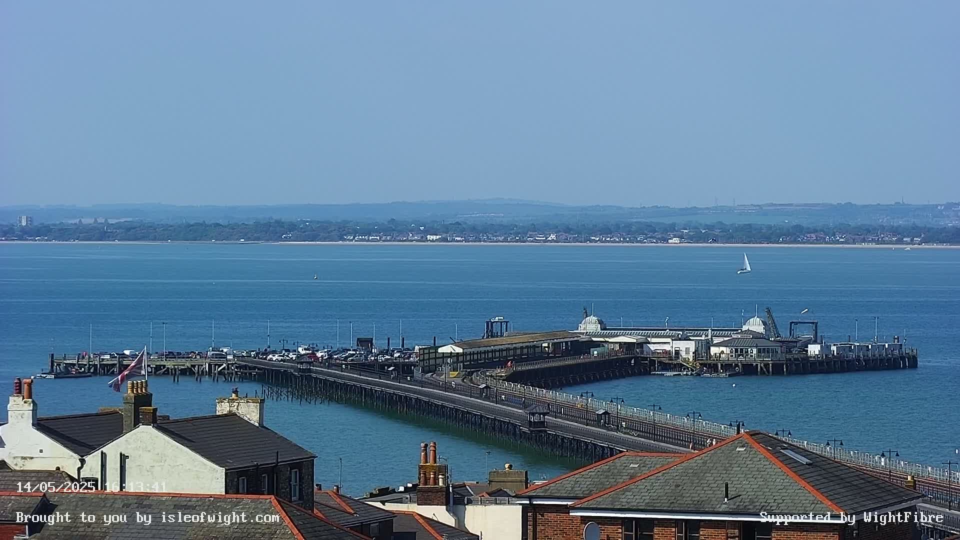 A long pier extends into a calm blue body of water on a sunny day, with a distant shoreline visible in the background.