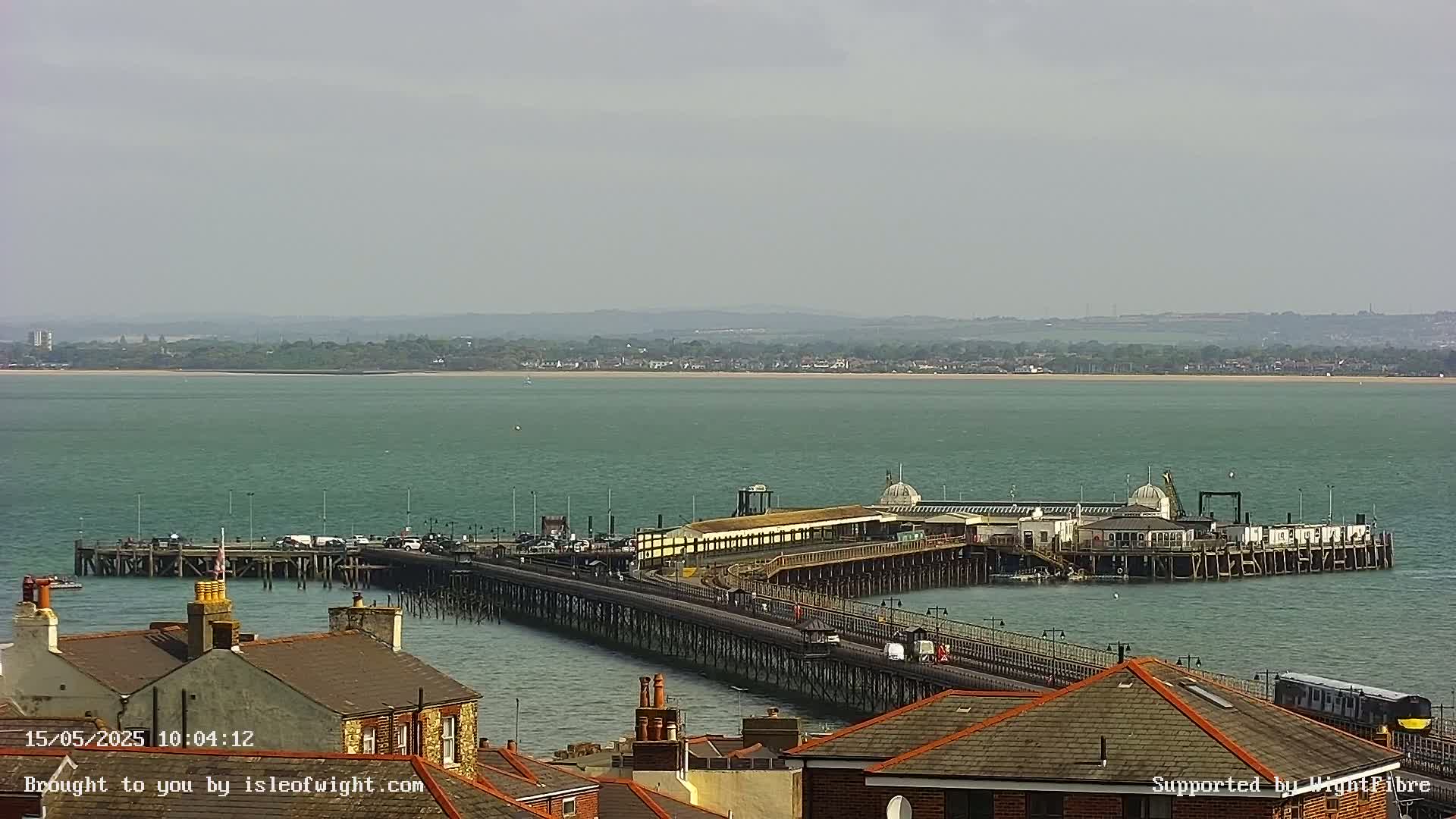 A long pier extends into a calm, teal sea under an overcast sky, with rooftops of buildings visible in the foreground.