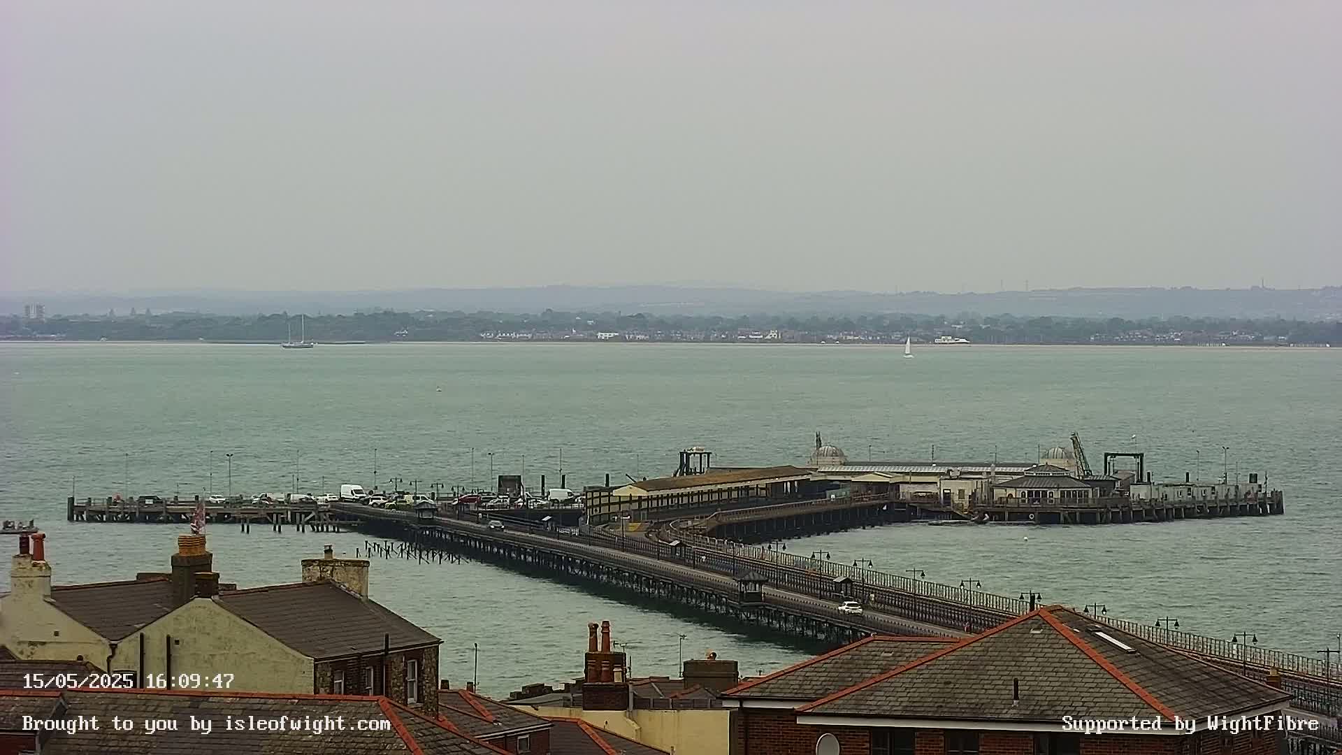 A long pier extends into a calm, grey sea, with a distant shoreline visible under an overcast sky.