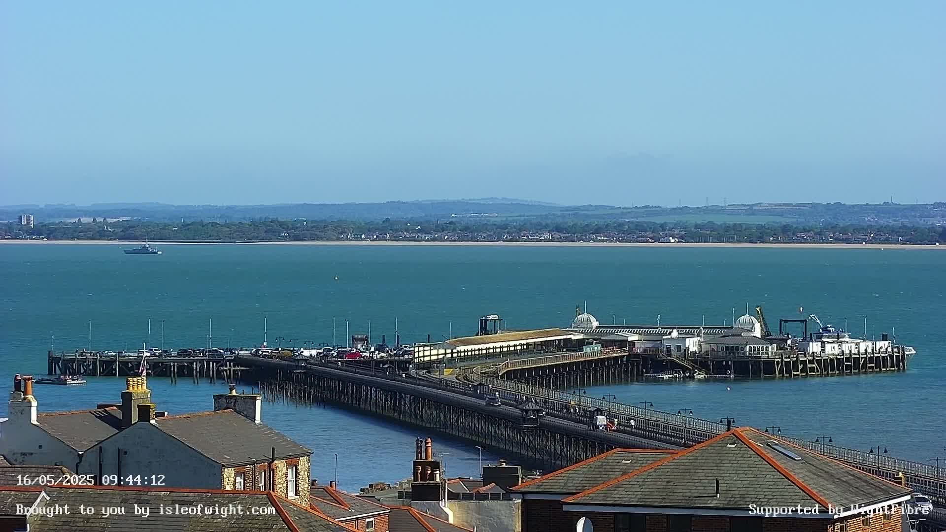 A long pier extends into a calm, teal-colored body of water on a sunny day, with a distant shoreline visible in the background.