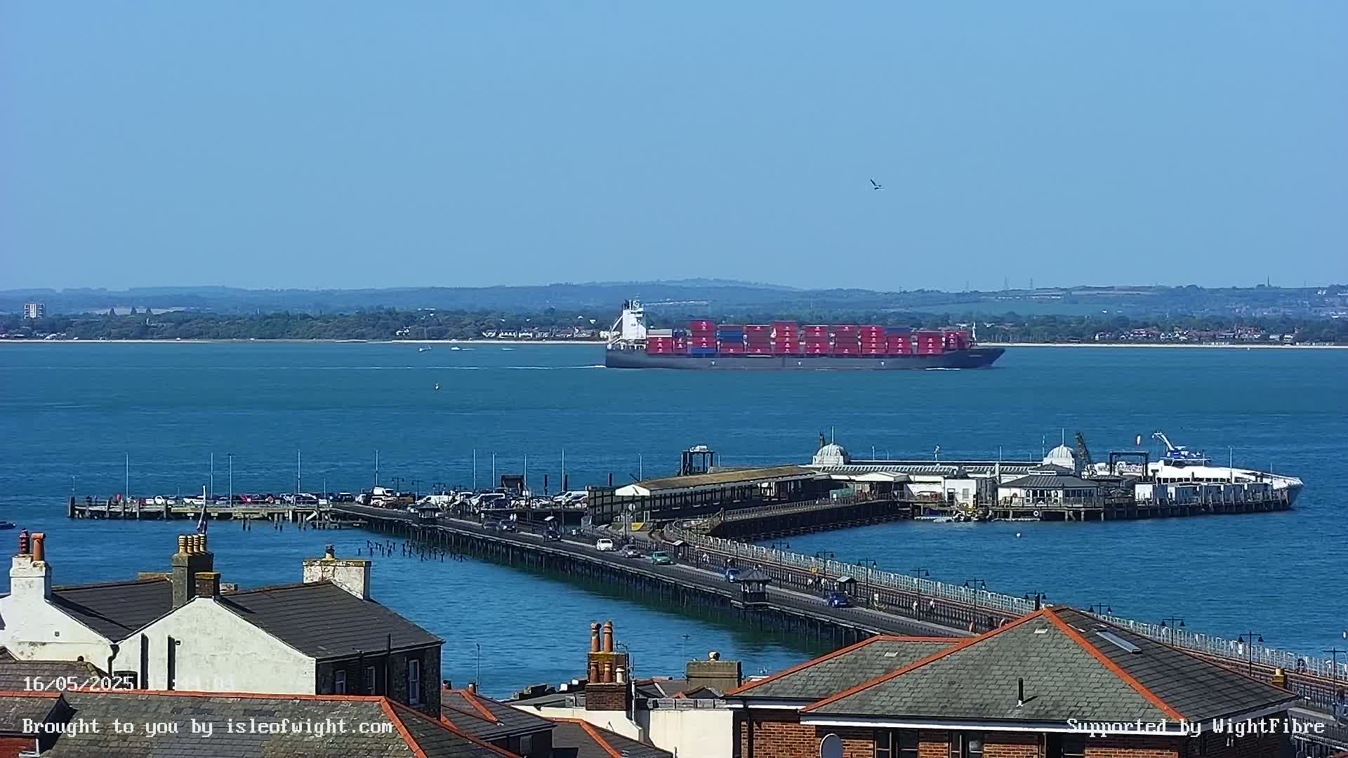 A large container ship sails past a pier extending from a town on a sunny day.