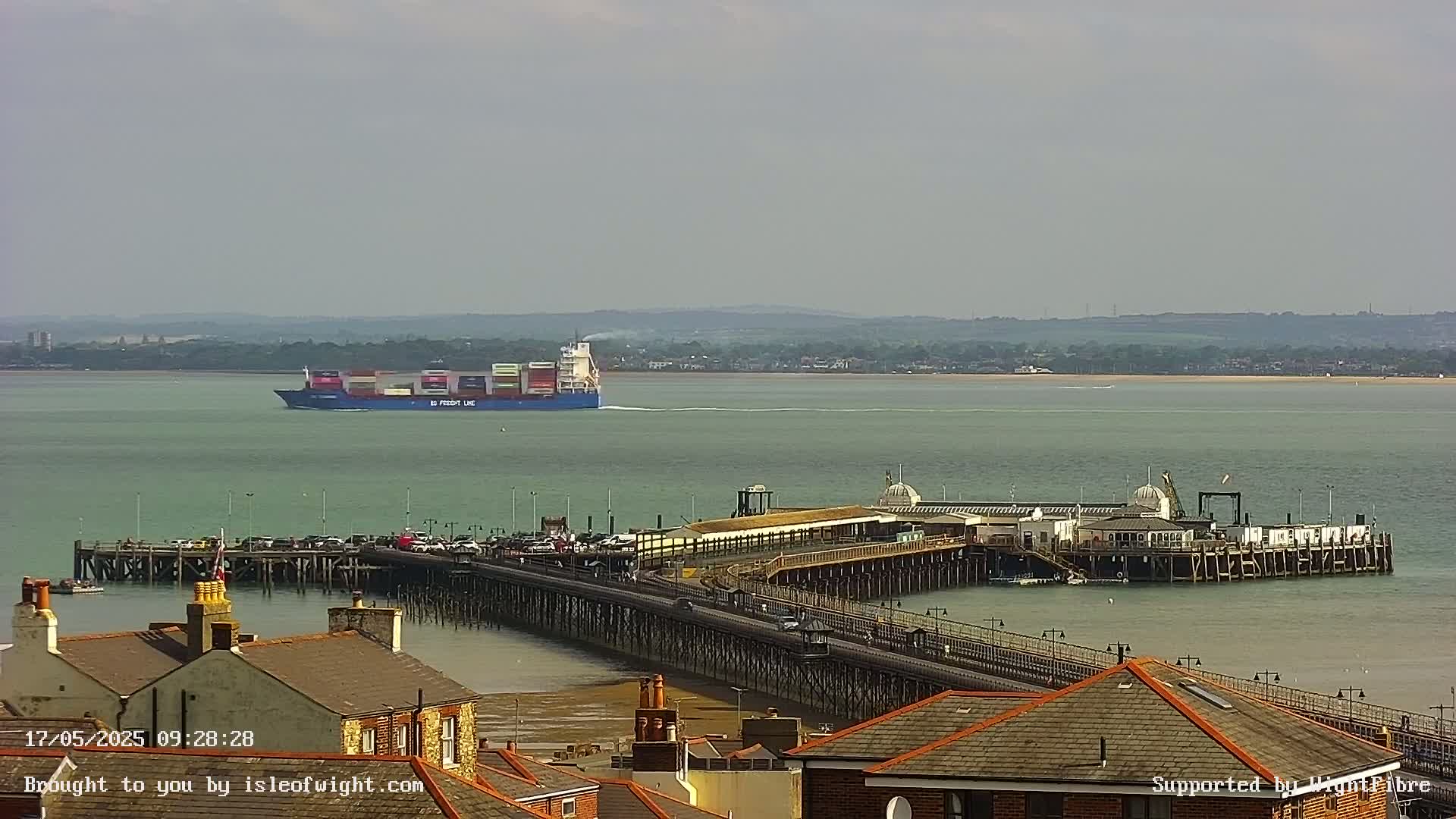A large container ship sails on calm water past a long pier with buildings and parked cars, viewed from an elevated position on a sunny day.