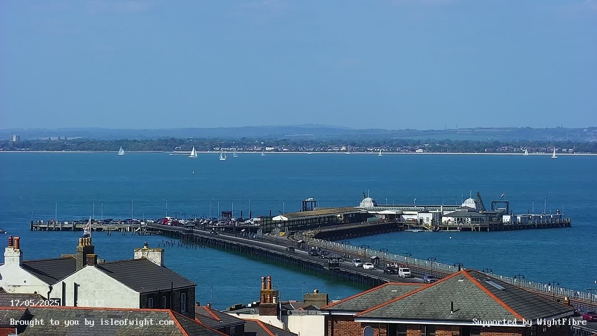 A long pier extends from a town into a calm, blue body of water on a sunny day, with several sailboats visible in the distance.