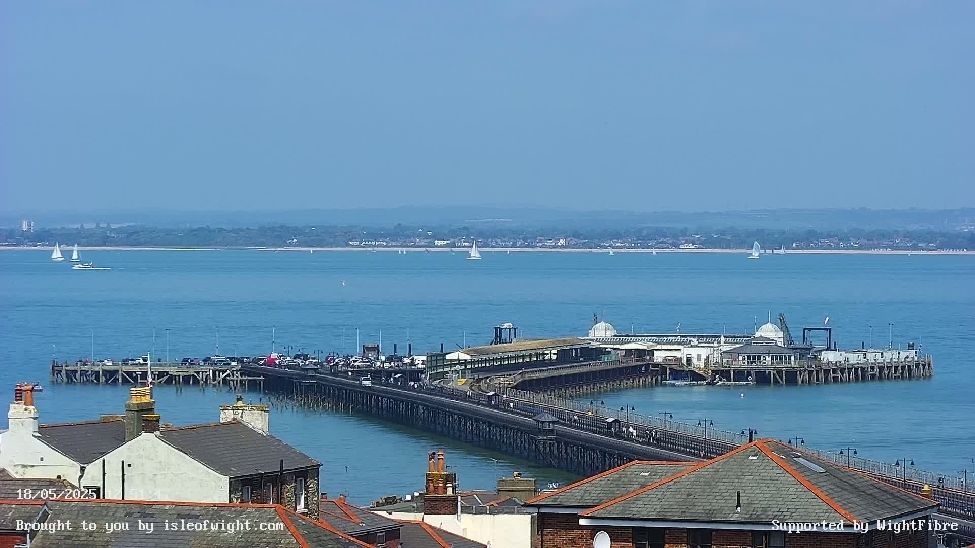 A long pier extends into a calm, blue sea on a sunny day, with several sailboats visible in the distance.