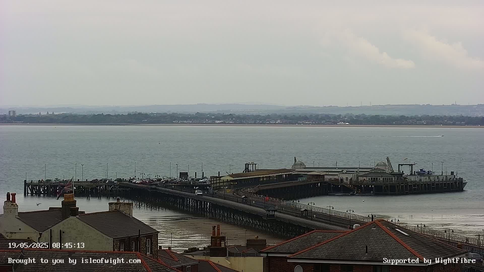 A long pier extends from a town's edge into a calm, gray-toned body of water, under an overcast sky.