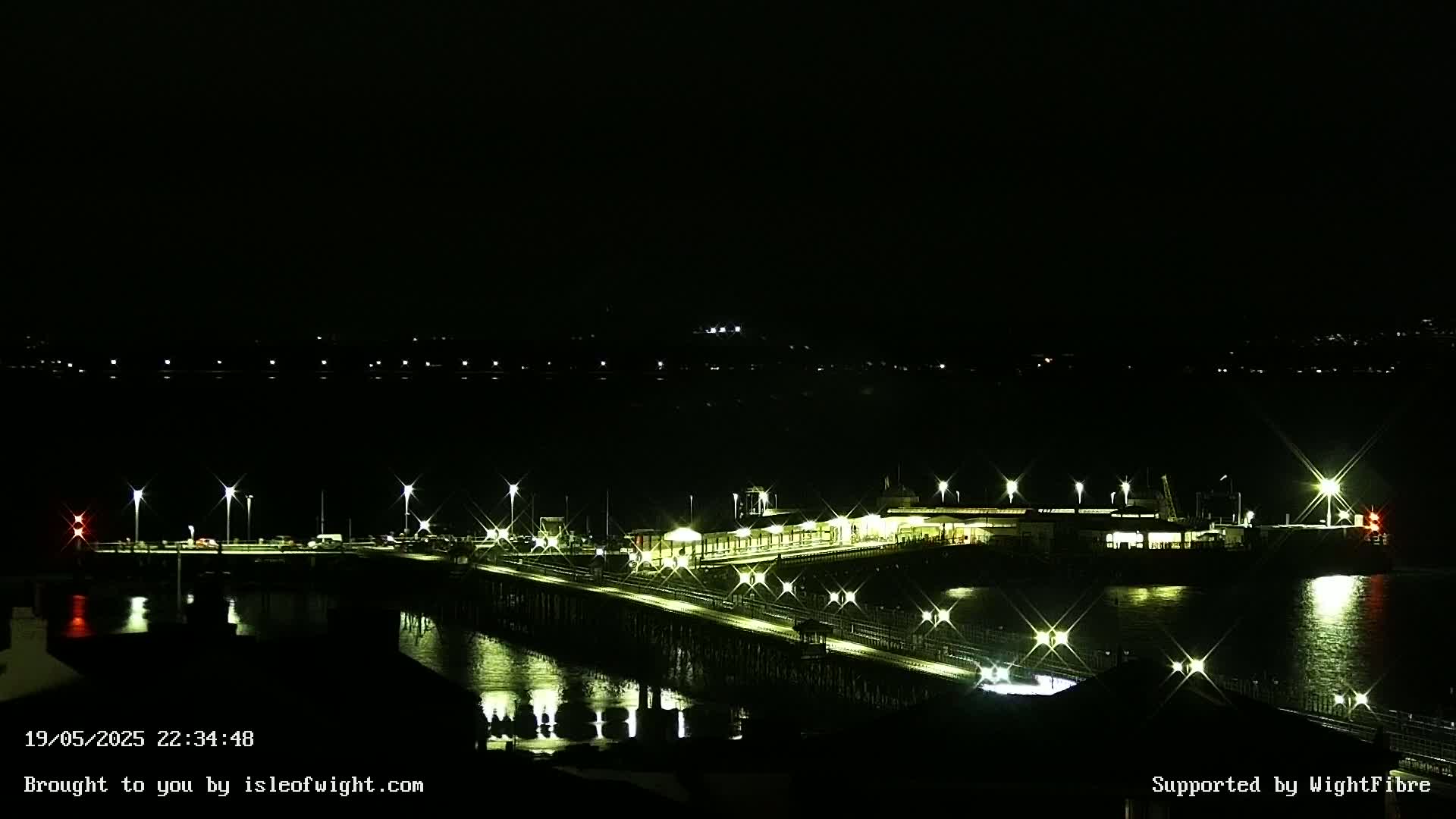 A brightly lit pier extends over dark water at night, with a building on the pier and distant lights on the shore.