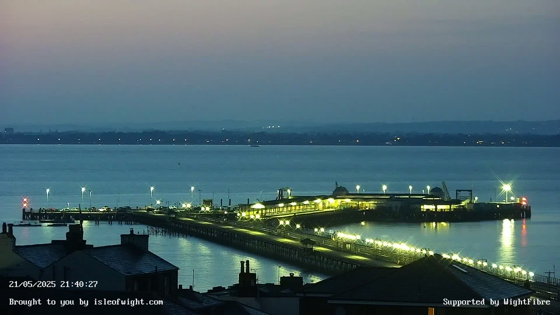 A long pier with lights illuminating its length extends from a shoreline into a calm, dark blue body of water under a twilight sky.