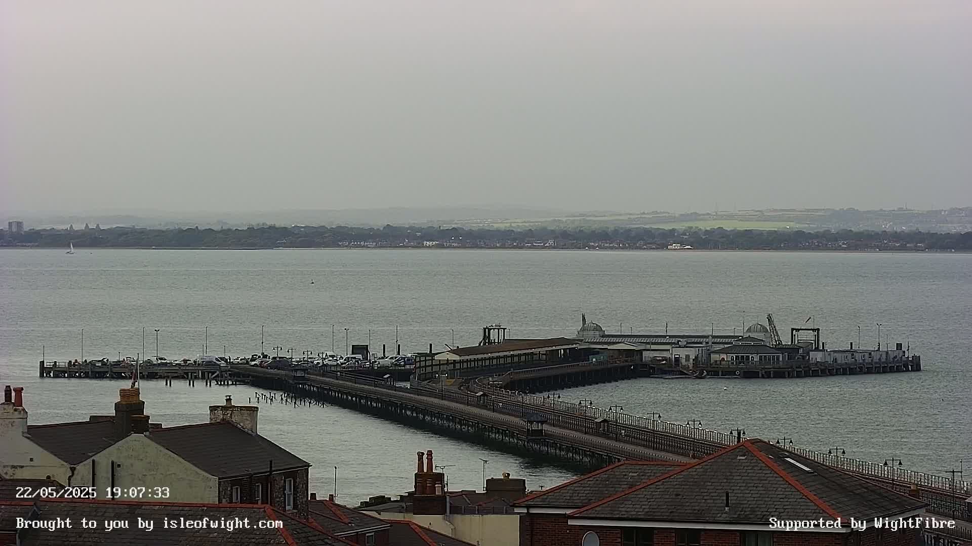 A long pier extends from a shoreline into a calm, gray body of water under an overcast sky.