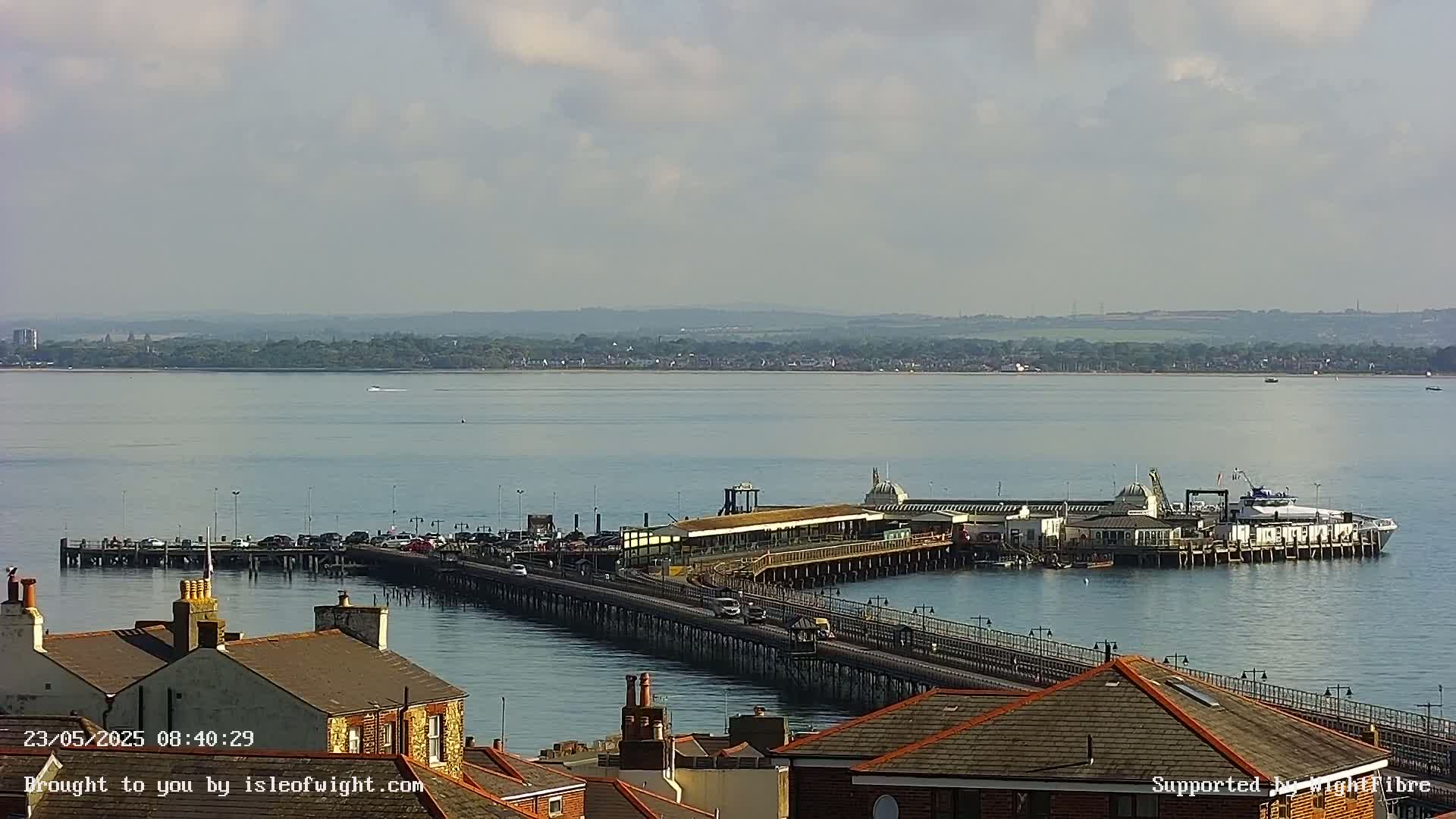 A long pier extends from a shoreline into a calm body of water under a partly cloudy sky, with a ferry docked at its end.