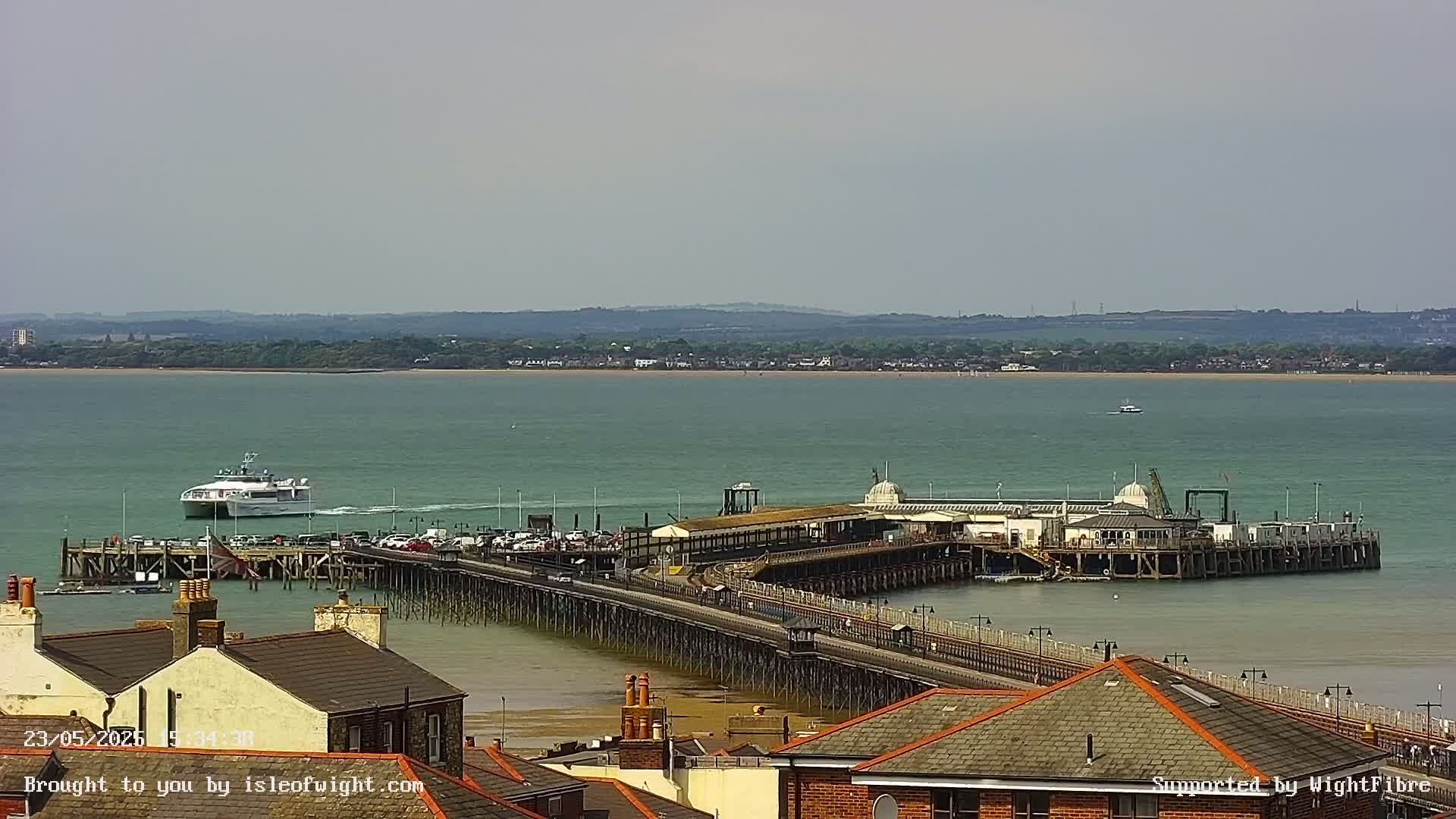 A long pier extends into a calm, green body of water, with a ferry approaching and buildings visible in the background under a mostly clear sky.
