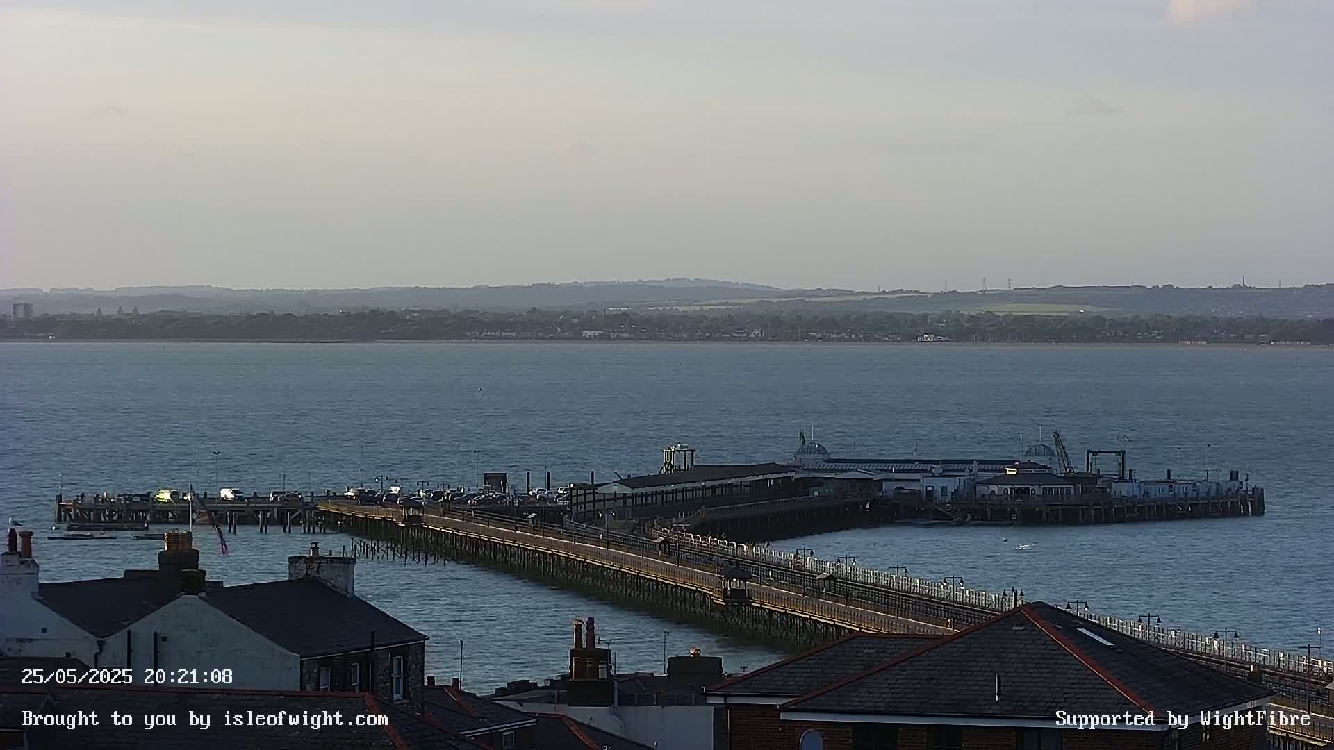 A long pier extends into a calm body of water under a hazy sky, with buildings visible in the foreground and a distant shoreline.