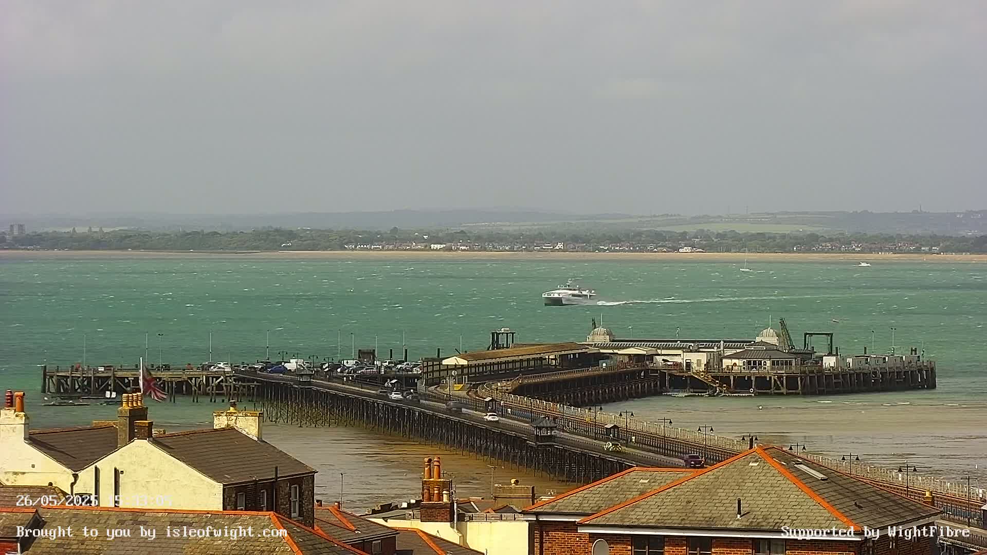 A long pier extends into a choppy, teal sea, with a ferry in the distance under a mostly cloudy sky.