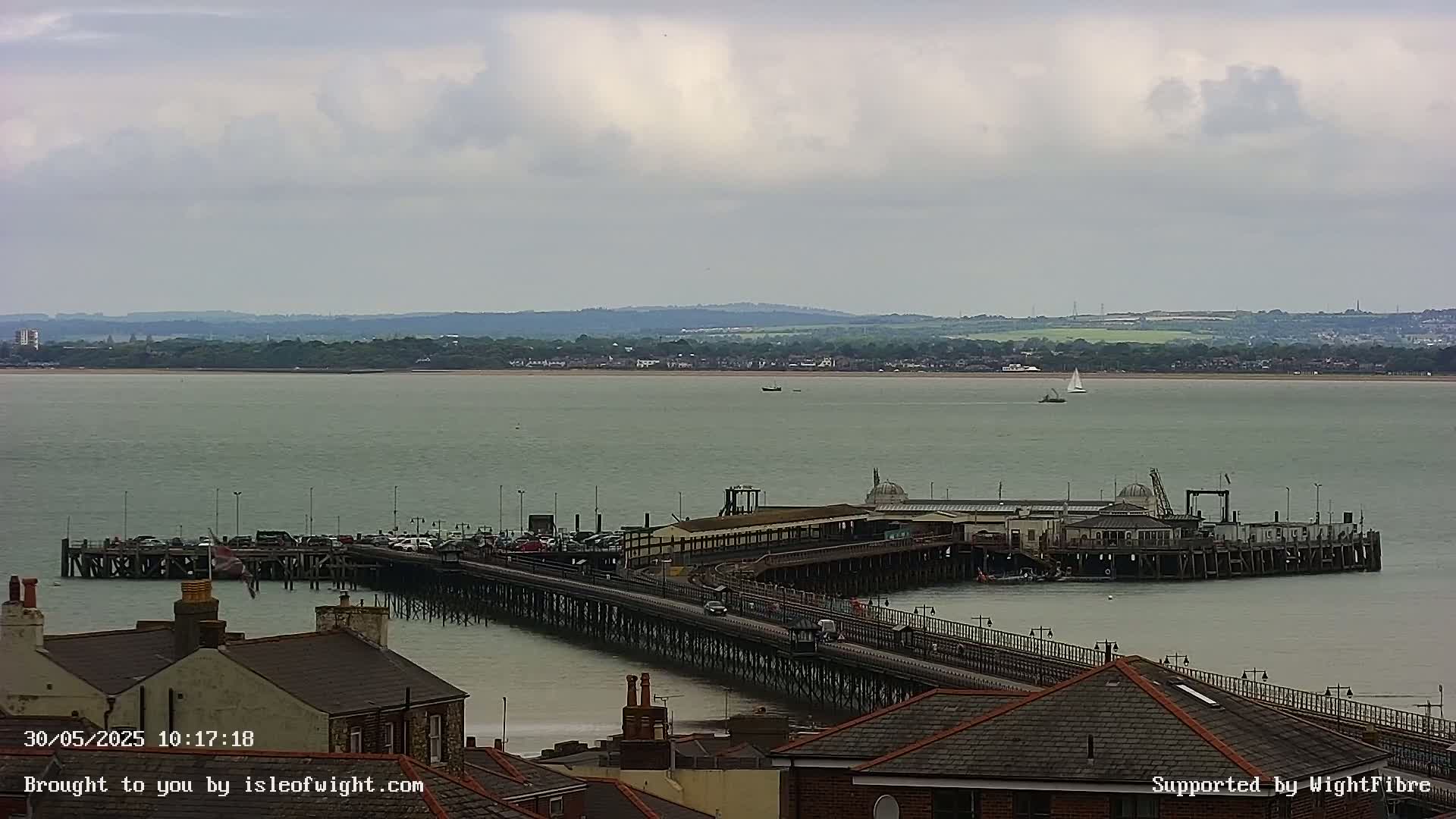 A long pier extends into a calm, grey-toned body of water under an overcast sky, with a distant shoreline visible.