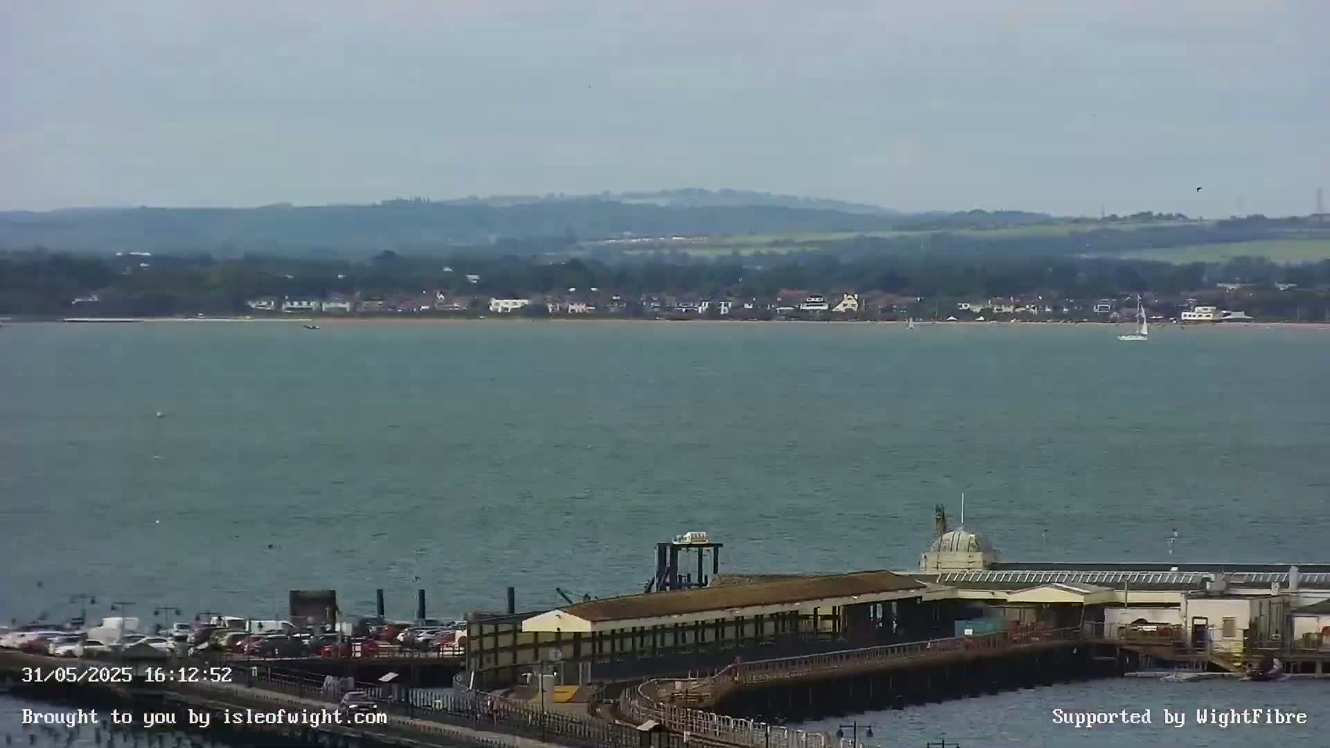 A long pier with a building at its end extends into a calm body of water, across from a distant shoreline with low-lying buildings and hills under an overcast sky.