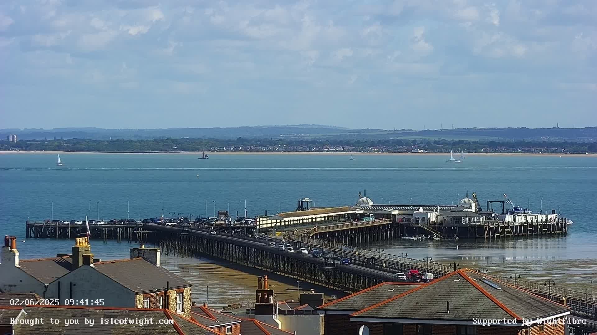 A long pier extends from a town into a calm, blue body of water on a partly sunny day, with several sailboats visible in the distance.