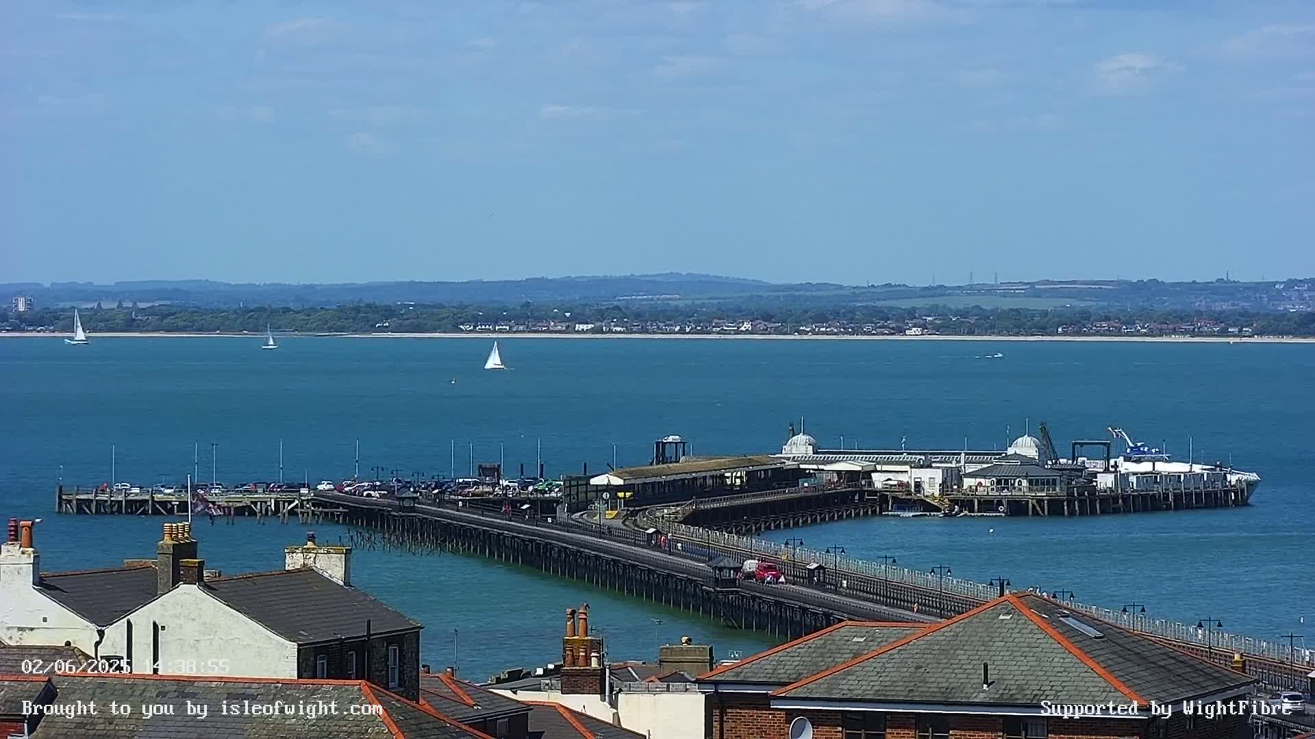 A long pier extends into a calm, blue body of water on a sunny day, with several sailboats visible in the distance and rooftops in the foreground.