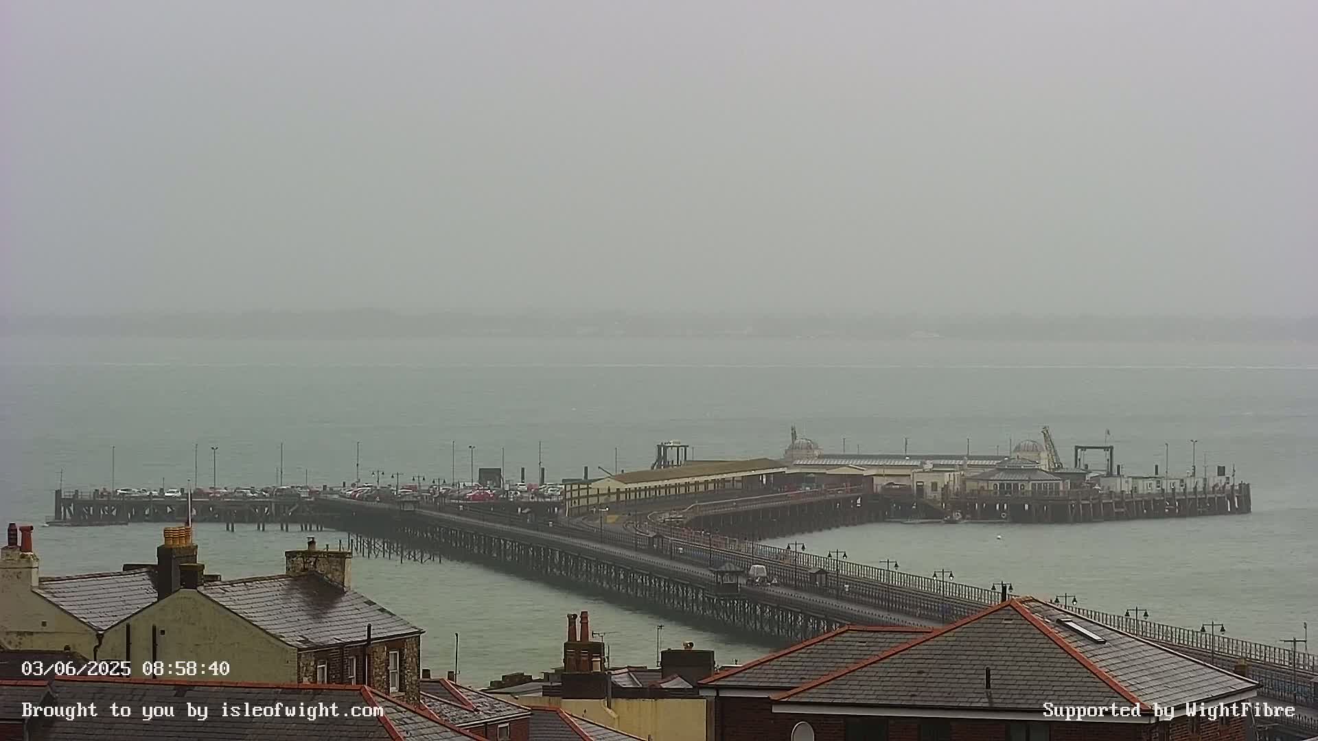 A long pier extends into a hazy, overcast sea, with rooftops visible in the foreground.