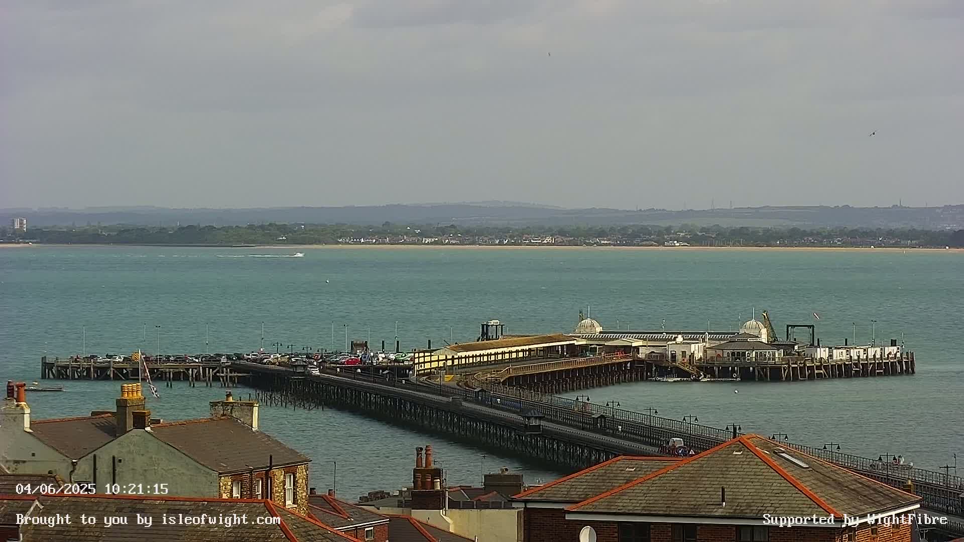 A long pier extends from a town into a calm, teal sea under a mostly cloudy sky.