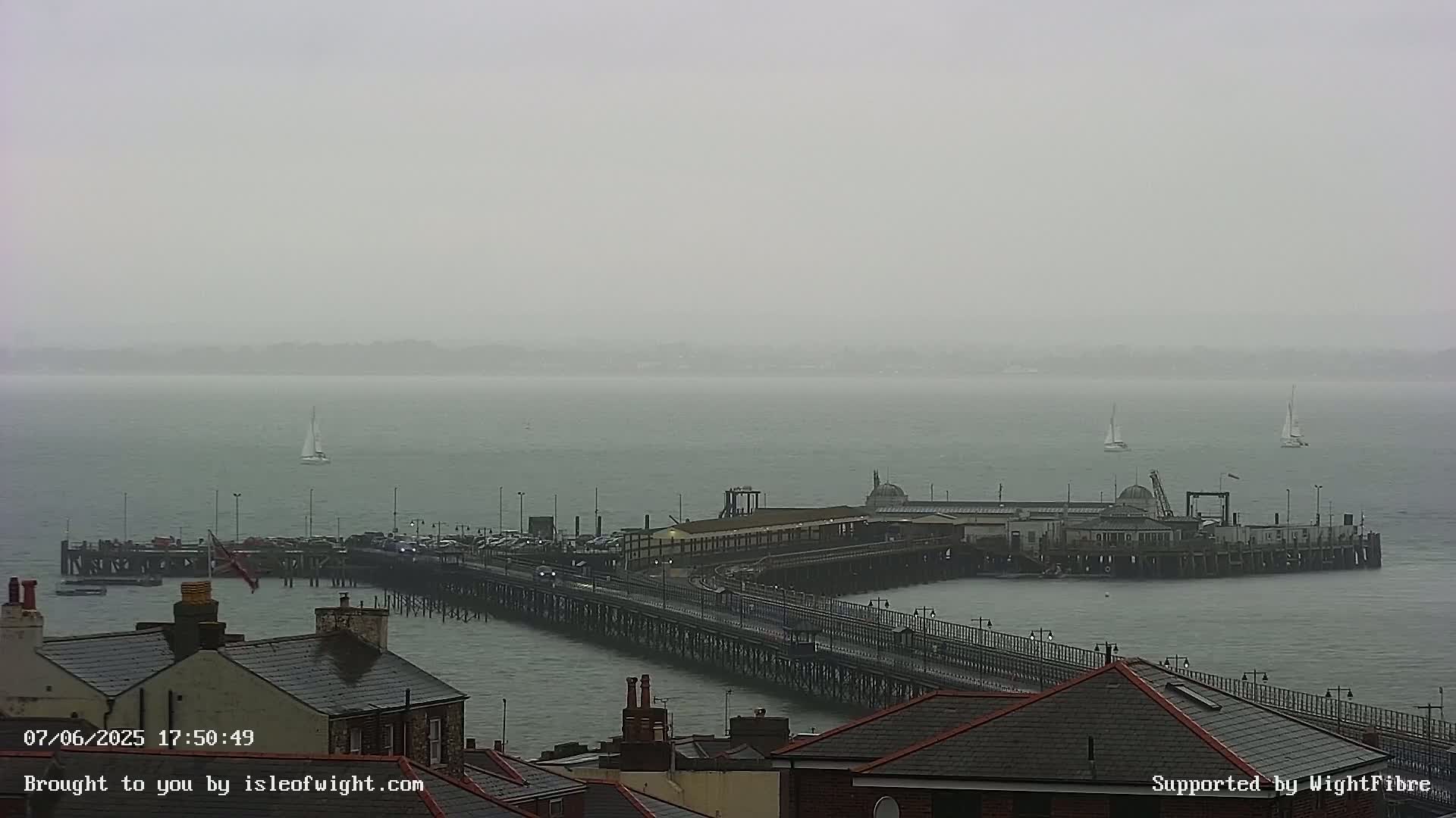 A long pier extends into a gray, overcast body of water, with several sailboats visible in the distance.