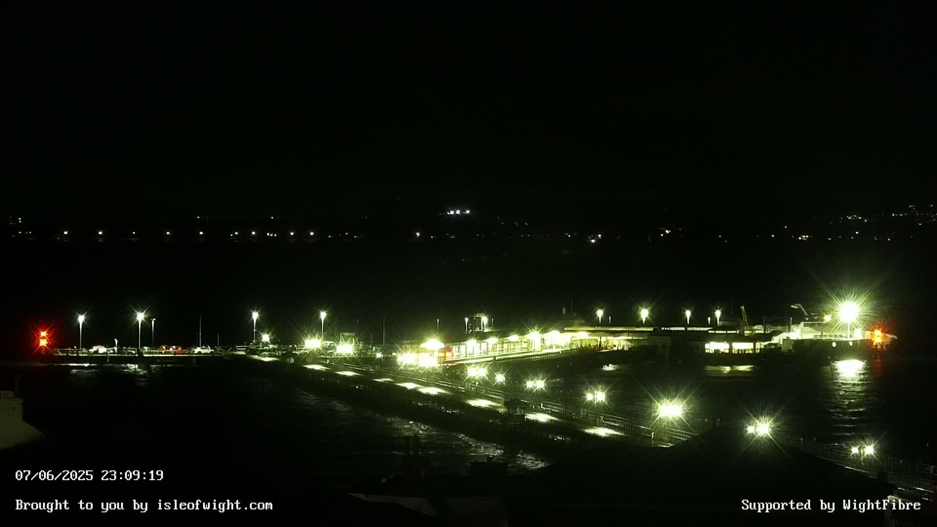 A brightly lit ferry terminal at night is seen from above, with dark water lapping at its edges.
