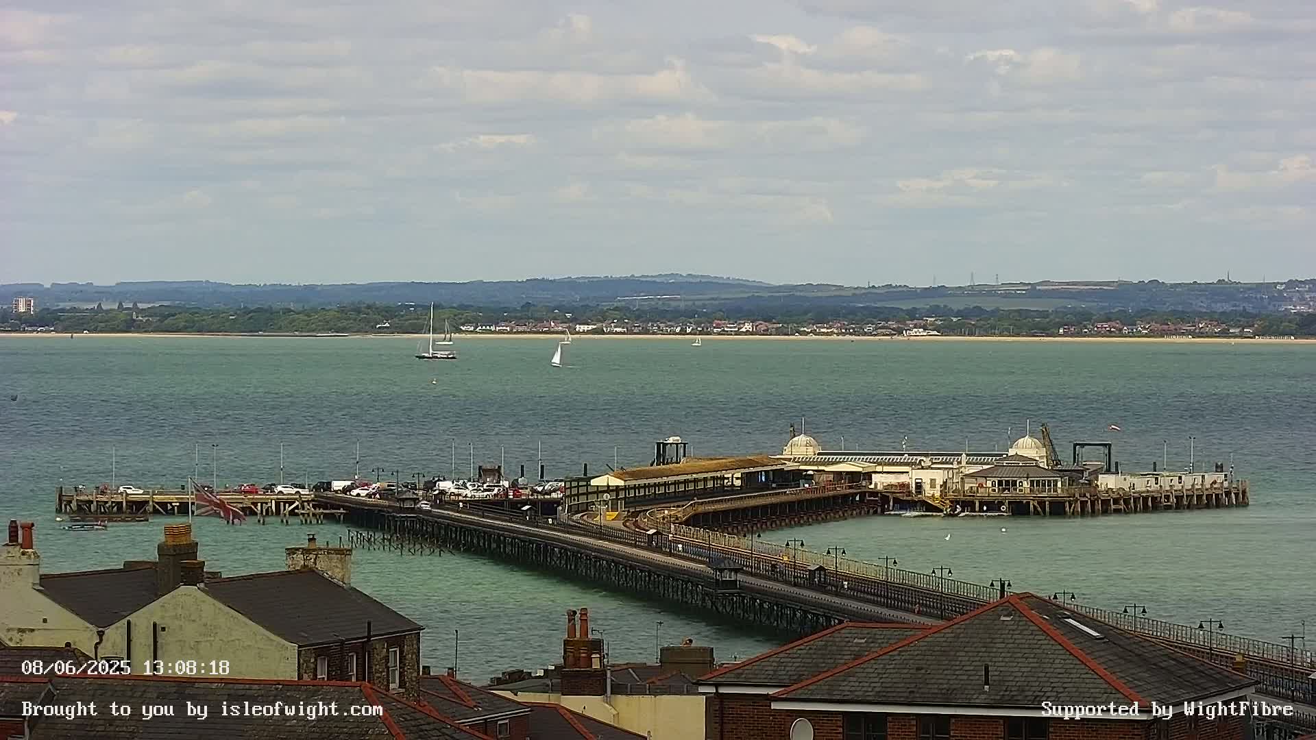 A long pier extends into a calm, green-blue sea under a mostly cloudy sky, with several sailboats visible in the distance and buildings visible in the foreground.