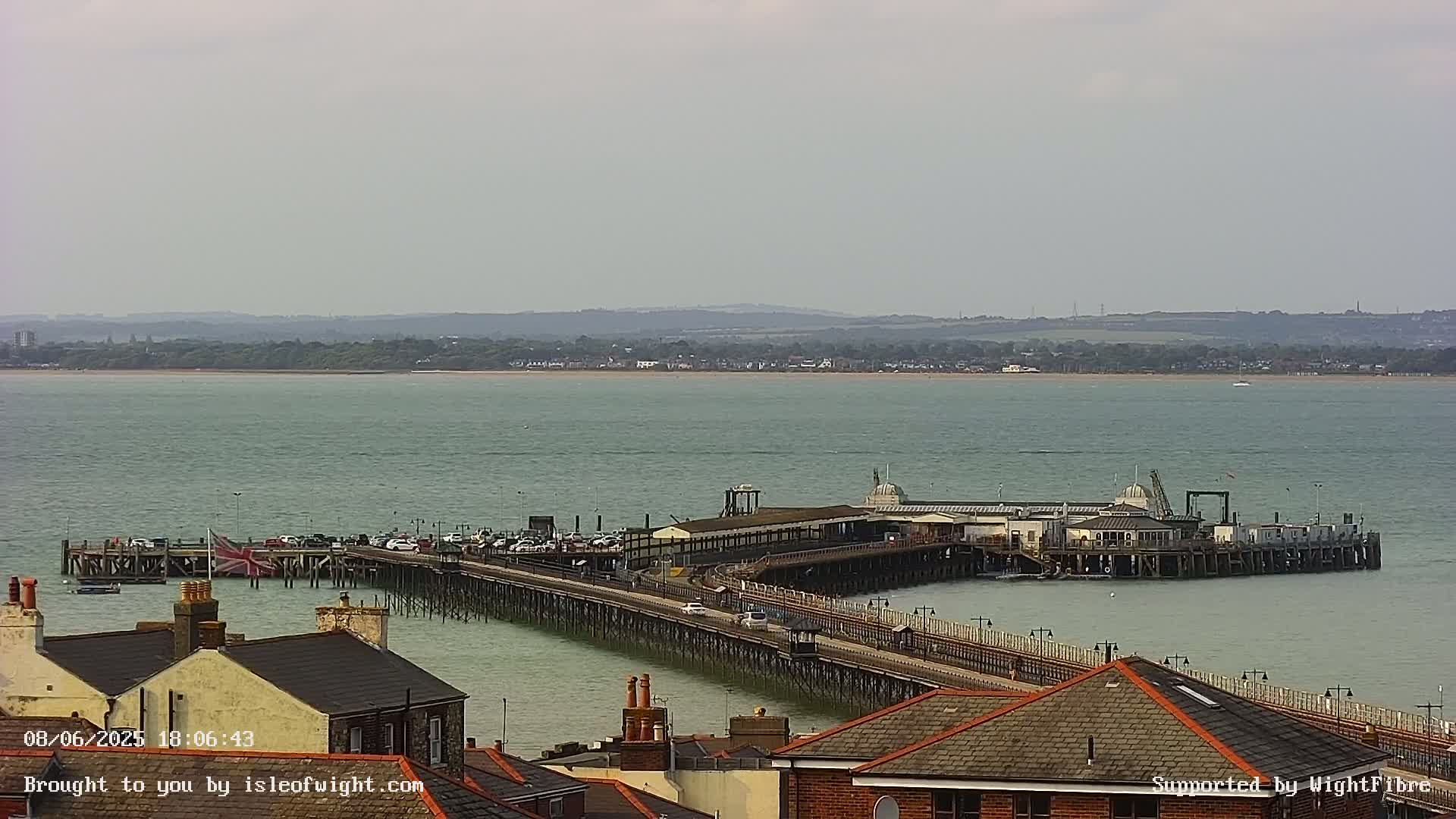A long pier extends into a calm, light-green sea under a mostly cloudy sky, with buildings and parked cars visible on the pier and in the foreground.