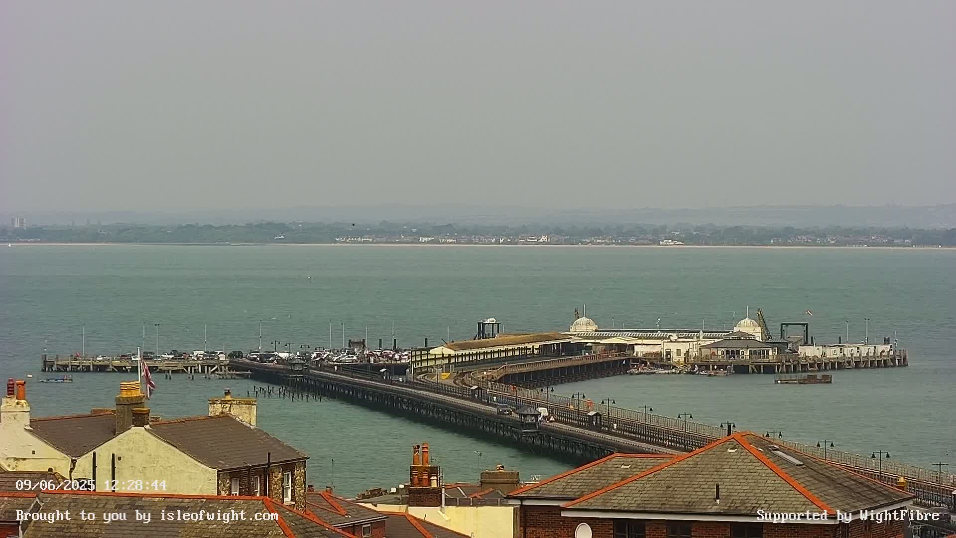 A long pier extends into a calm, hazy sea, with buildings and rooftops visible in the foreground.