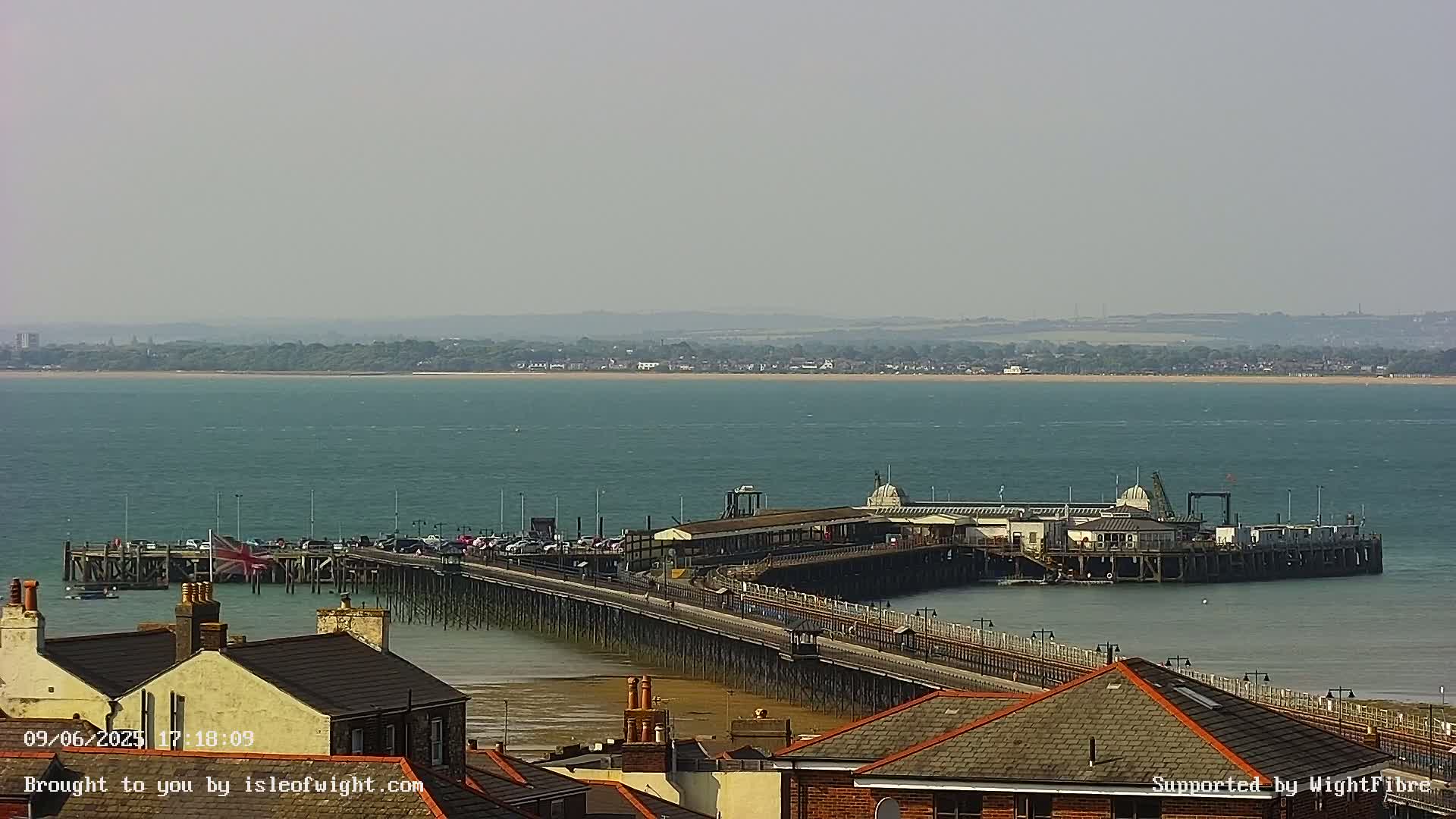 A long pier extends from a shoreline into a calm, teal sea under a hazy sky.