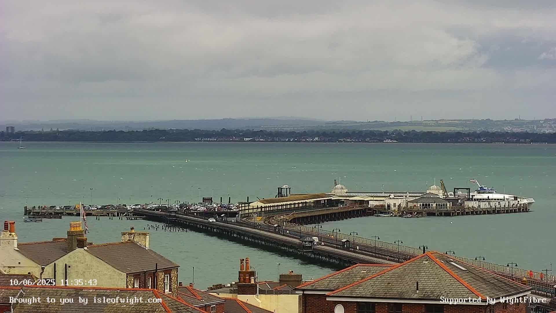 A long pier extends from a shoreline into a calm, green body of water under a cloudy sky, with buildings and rooftops visible in the foreground.