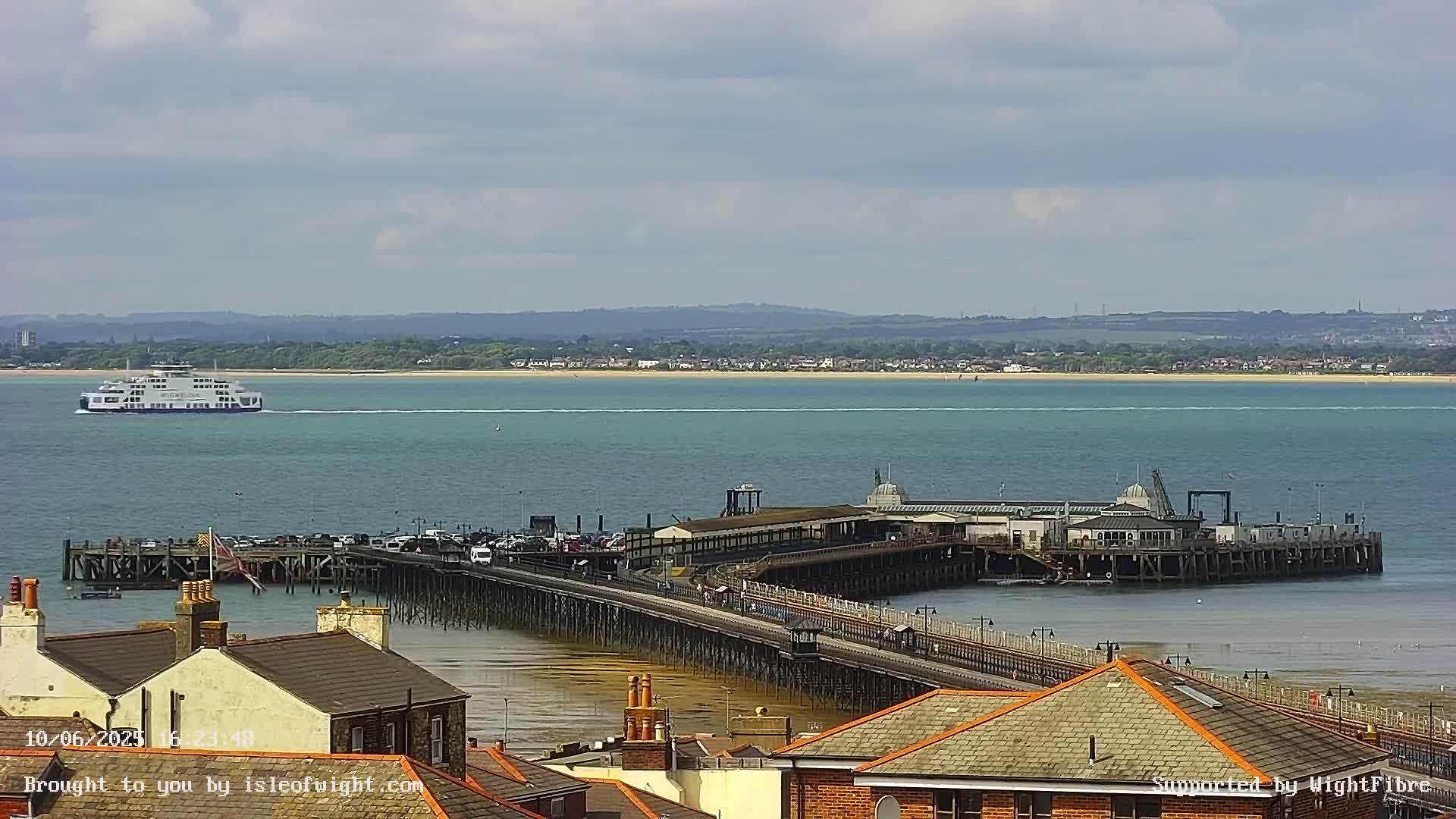 A long pier extends into a calm, teal-colored body of water, with a ferry visible in the distance under a partly cloudy sky.