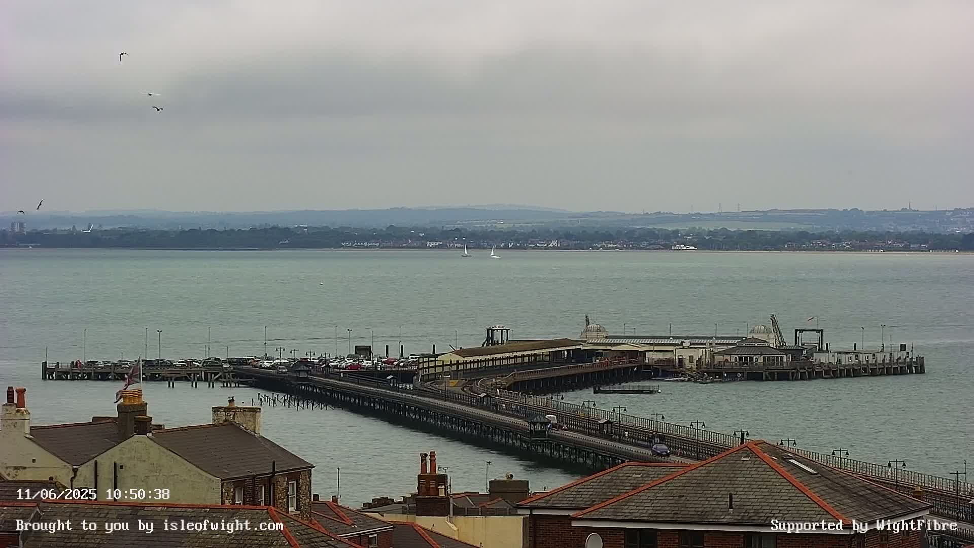 A long pier extends into a calm, grey-sky sea, with rooftops and a distant shoreline visible.