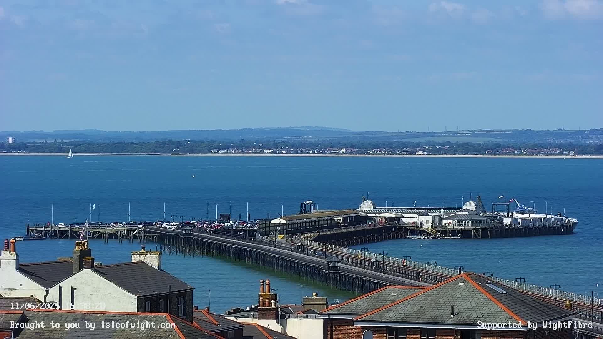 A long pier extends into a calm blue body of water on a sunny day, with a distant shoreline visible in the background.