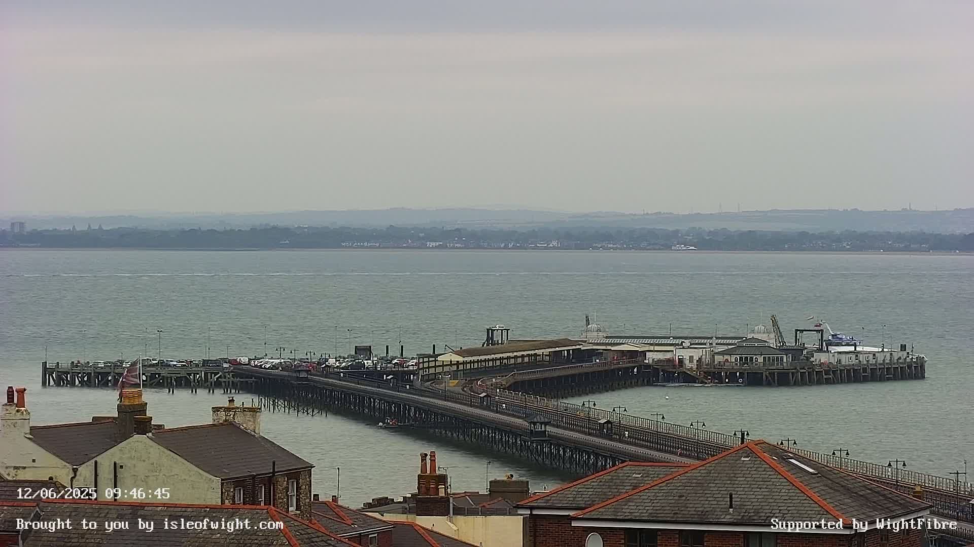 A long pier extends into a calm, gray body of water under an overcast sky, with buildings and rooftops visible in the foreground.