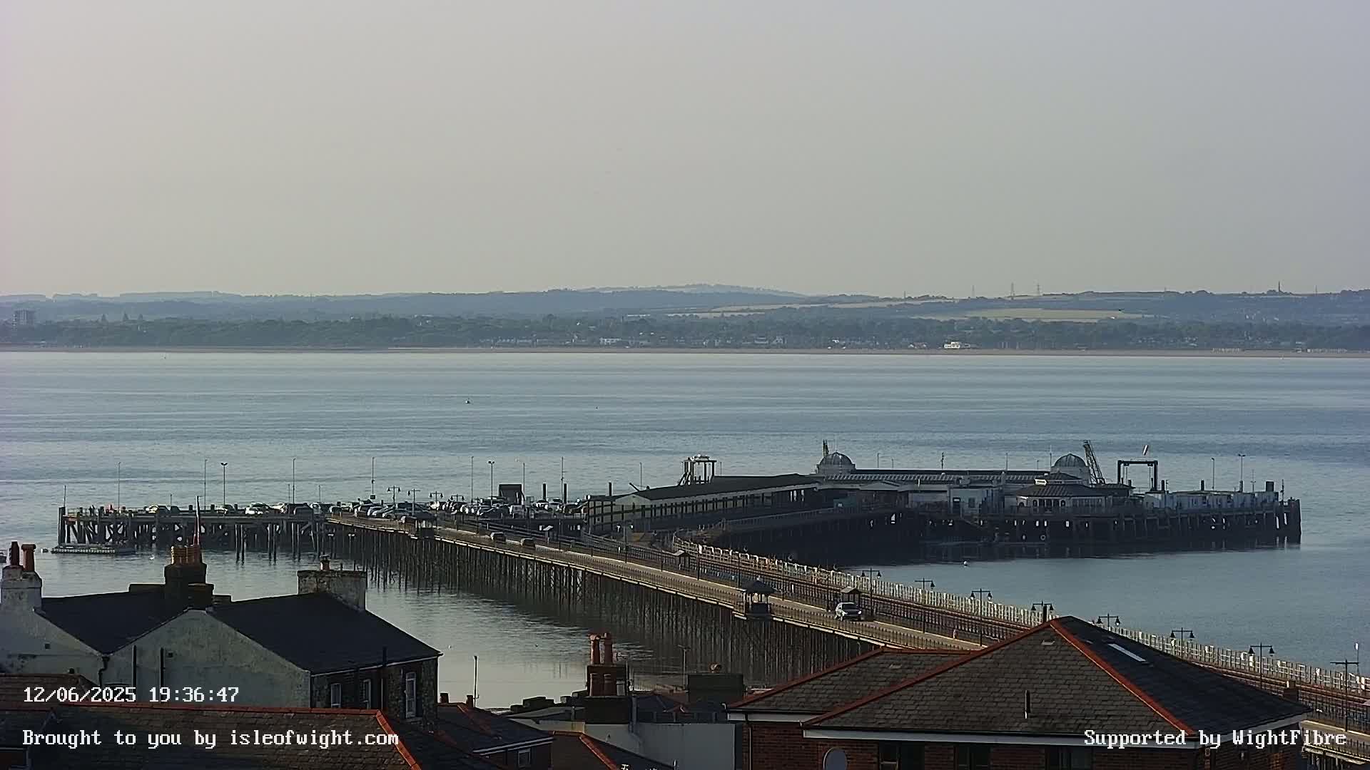 A long pier extends into a calm body of water under a hazy sky, with buildings and rooftops visible in the foreground.