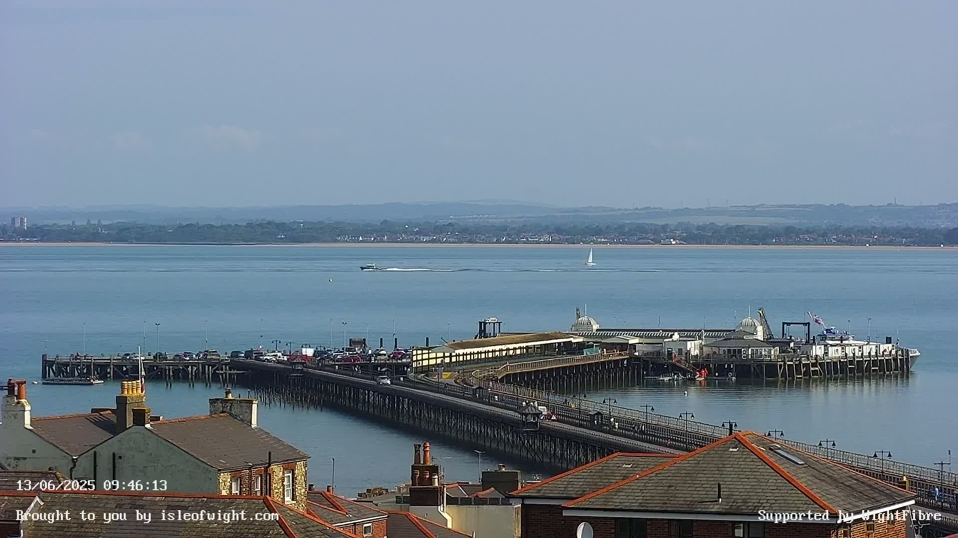 A long pier extends into a calm, blue body of water on a sunny day, with a distant shoreline visible in the background and rooftops in the foreground.
