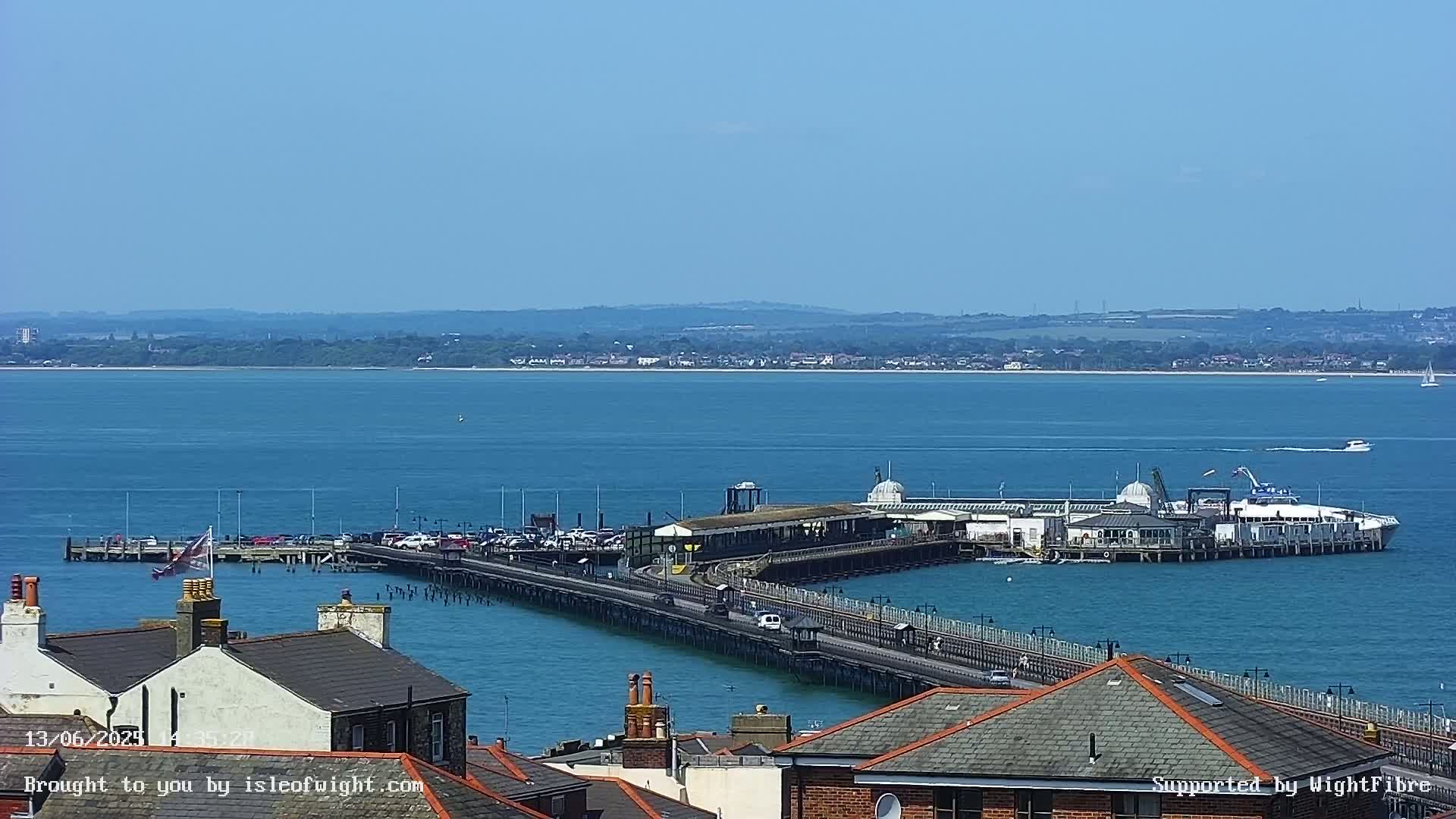 A long pier extends into a calm, blue body of water on a sunny day, with a building complex at its end and town rooftops visible in the foreground.