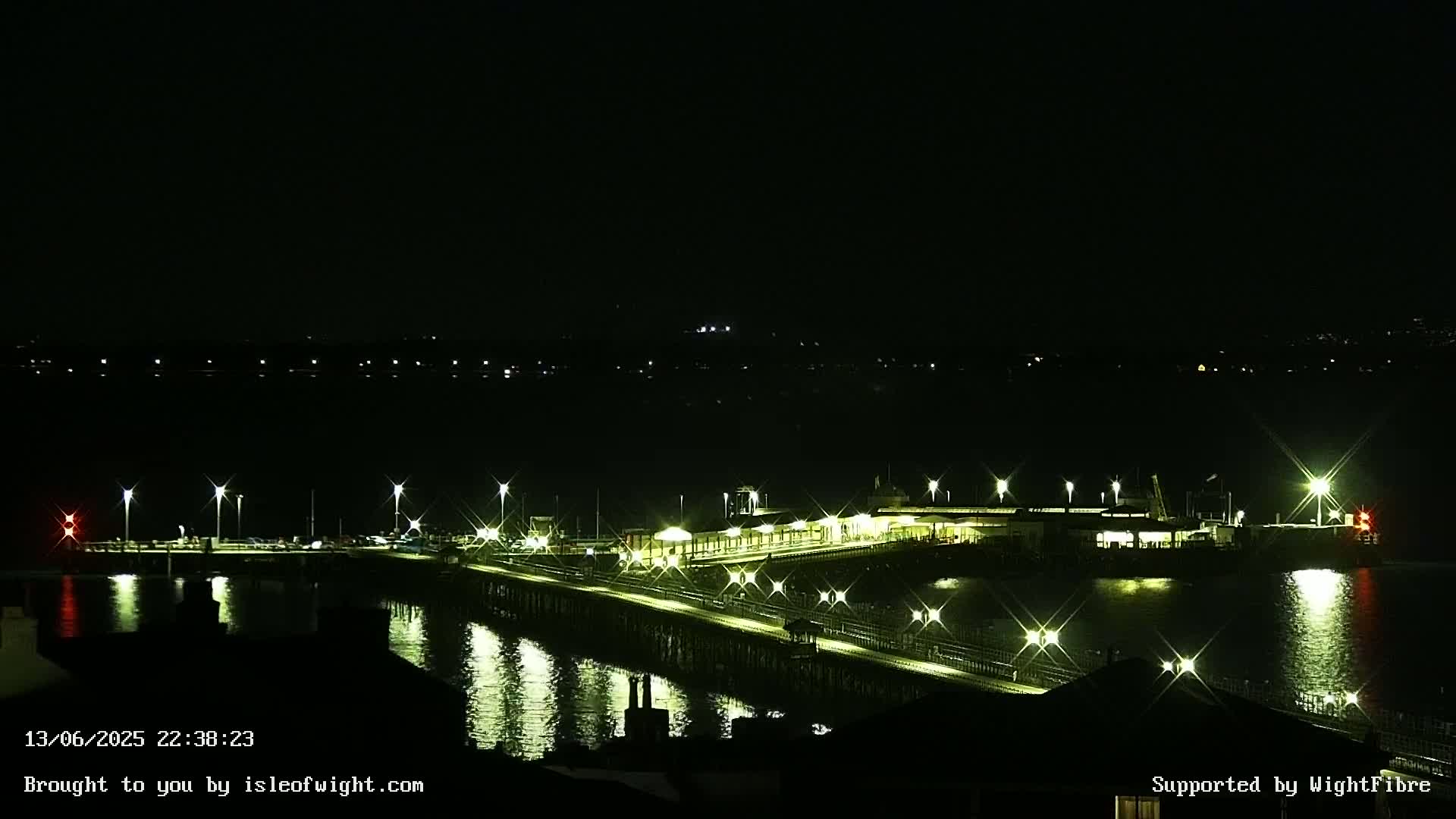 A brightly lit pier extends over dark water at night, with lights illuminating the shoreline in the distance.
