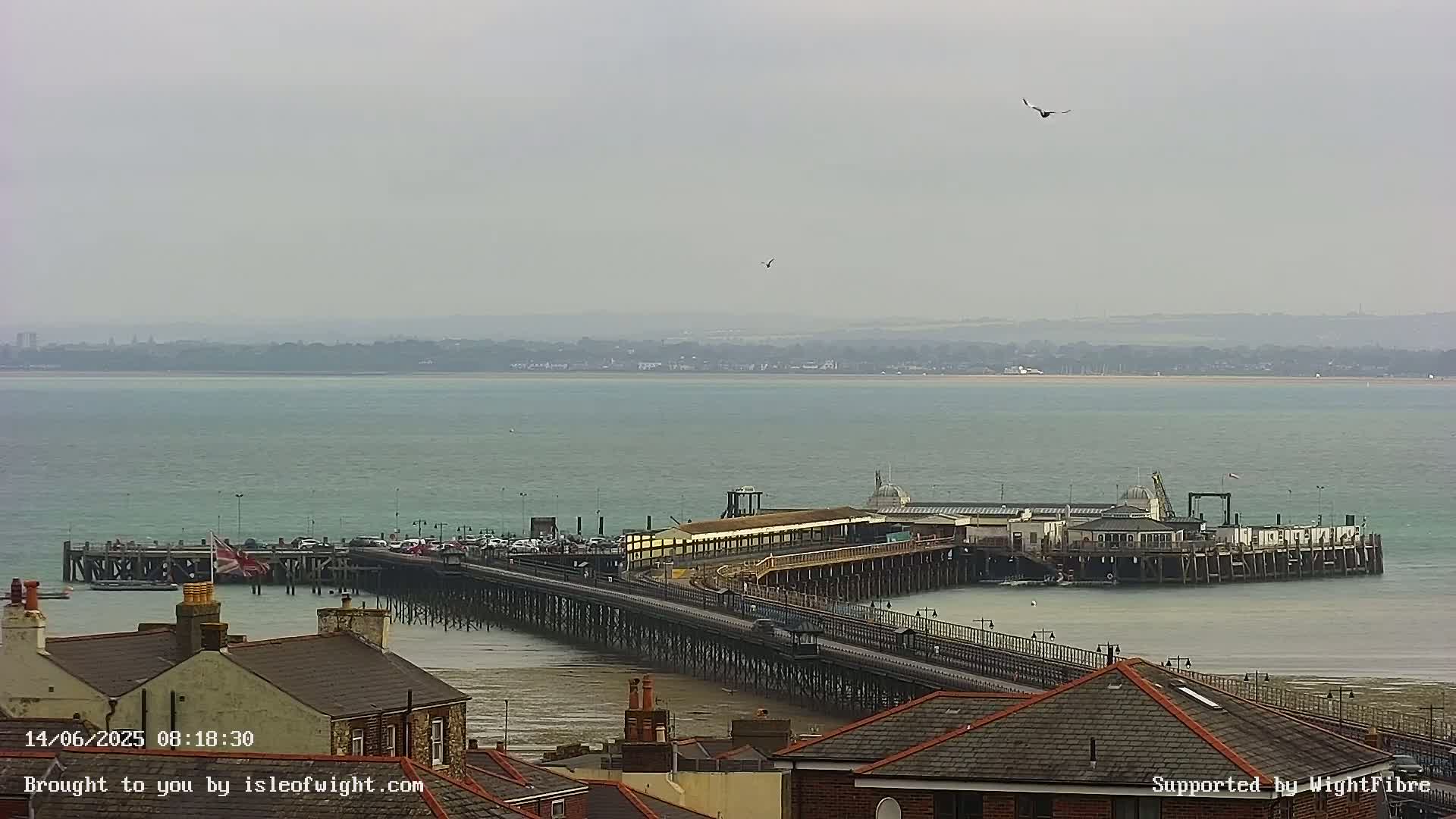A long pier extends into a calm, light-grey sea under an overcast sky, with rooftops of buildings visible in the foreground.